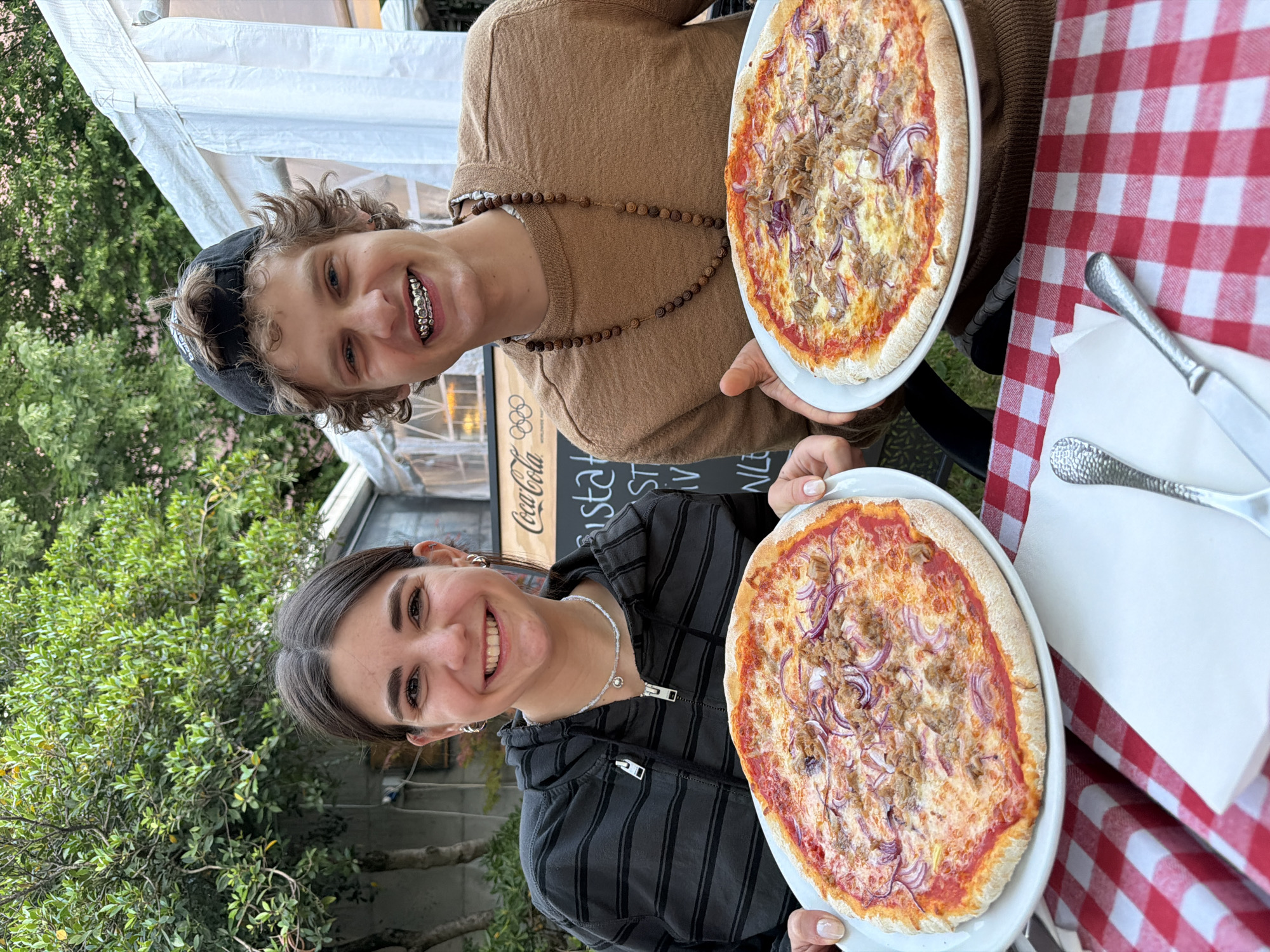 This outdoor scene in Milan, Italy, captures two young individuals happily posing with their pizzas. A male, with curly brown hair under a black cap, smiles, revealing metallic dental grills on his upper teeth. He wears a brown long-sleeved shirt and a long wooden beaded necklace. Next to him, a female with dark hair and a silver necklace smiles at the camera, dressed in a dark striped hooded jacket.
Each holds a freshly prepared pizza, identical in appearance, topped with red sauce, cheese, red onions, and shredded meat, likely tuna. They are positioned at a table covered with a classic red and white checkered tablecloth, with a set of cutlery (fork and knife) resting on a white napkin.
In the background, green trees and a white tent or awning structure are visible. A dark sign displays the Coca-Cola logo, Olympic rings, and partially visible white text that begins with "STA". The natural lighting suggests it is daytime. Both subjects appear cheerful, enjoying their meal in a casual, outdoor dining setting.