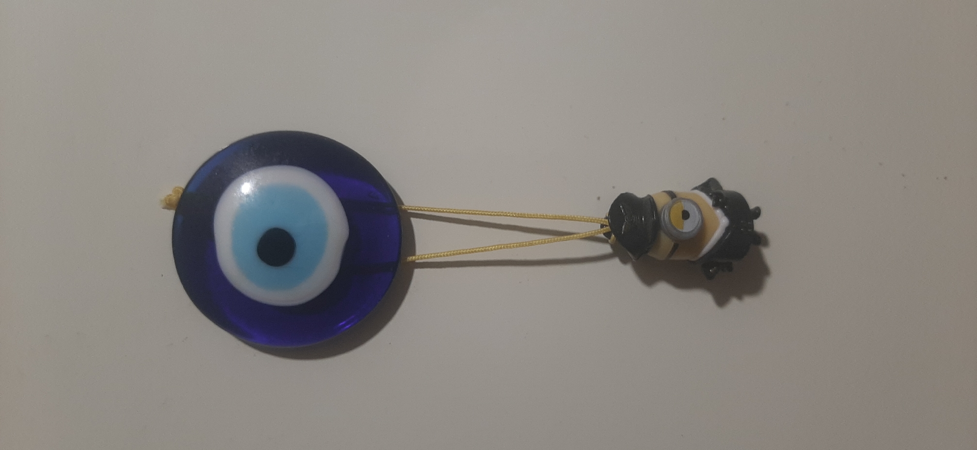 A close-up, vertically oriented shot shows a blue and white evil eye charm hanging against a plain, light-colored wall. Below the charm, a small, yellow Minion figurine, dressed in a black suit and hat, dangles from a yellow string. The Minion is facing slightly to the left. The background is uniformly lit, with minimal shadows, suggesting indoor lighting. No specific location in Yessentukskaya, Russia, can be determined from the image.