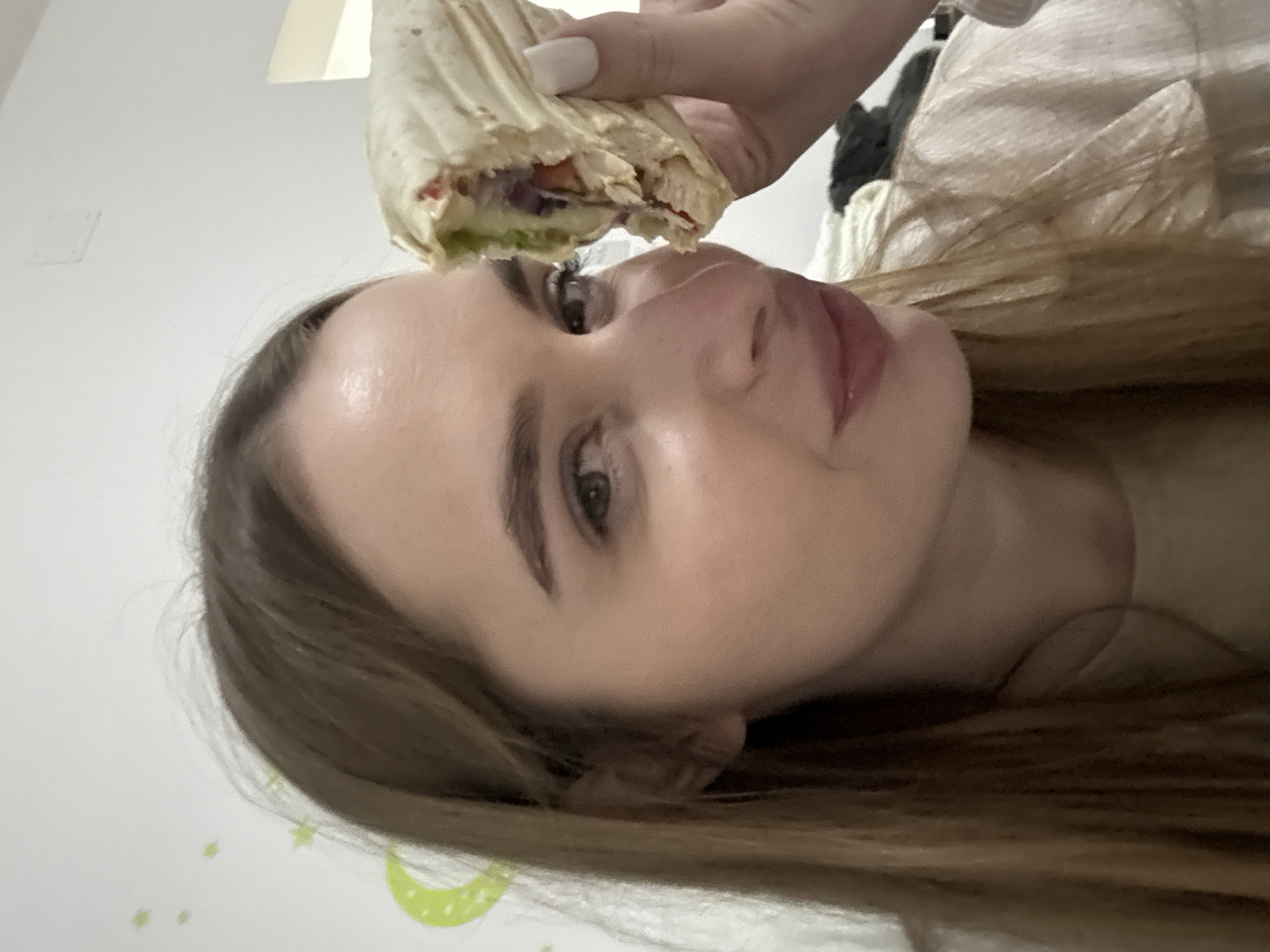 The image depicts a young woman in an indoor setting in Budva, Montenegro. She has long, light brown hair and is looking directly at the camera with a subtle expression. Her left hand, displaying white nail polish, holds a partially consumed wrap or shawarma close to her face. The visible contents of the wrap include chunks of meat, red cabbage, and green vegetables. The background is a plain white wall, with faint green star and crescent moon decals visible on the upper left side. The lighting is uniform, suggesting an indoor environment.