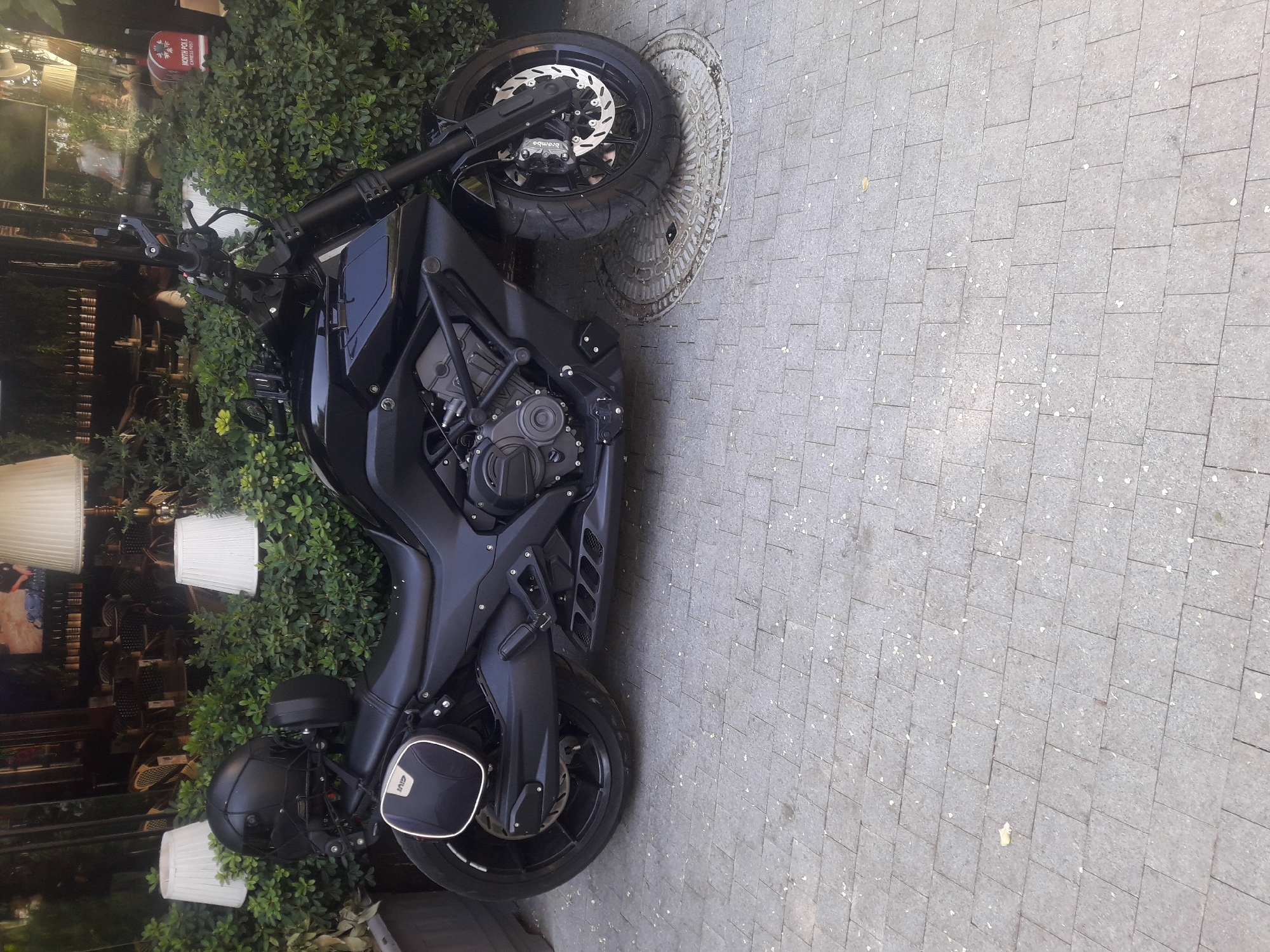 A black motorcycle with a black helmet on its seat is parked on a paved sidewalk in front of a restaurant with outdoor seating. The motorcycle has a black frame and black wheels, with a single saddlebag on the rear left side. The helmet is also black and appears to be a full-face helmet. The restaurant has large windows and several tables and chairs visible through them. There are also several large potted plants and lamps scattered around the outdoor seating area. The sidewalk is made of gray cobblestones, and there are a few small white petals scattered on the ground. The scene is well-lit, suggesting it is daytime.