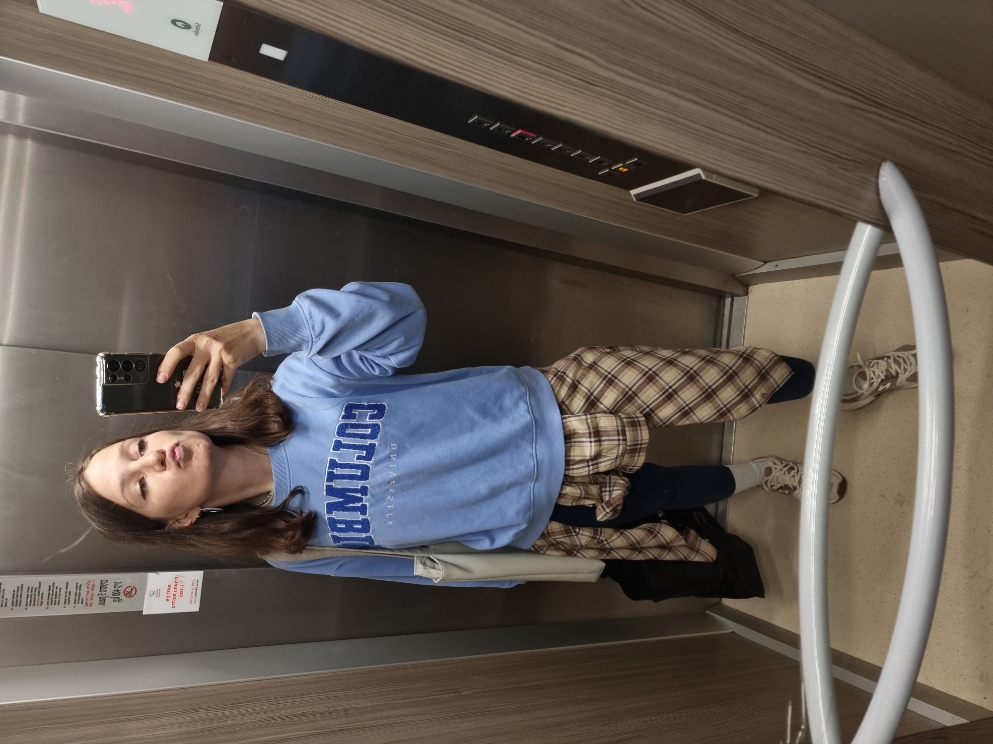 The image shows a young woman taking a selfie in what appears to be an elevator. She is wearing a light blue sweatshirt with "COLUMBIA" written on it in large, dark blue letters, a plaid shirt tied around her waist, dark pants, and light-colored sneakers. She is holding a smartphone with a black case. In the background, there is an elevator control panel with buttons and a digital display. There are also signs in Hebrew. The location is likely Ramat Gan, Israel, based on the Hebrew signage.
