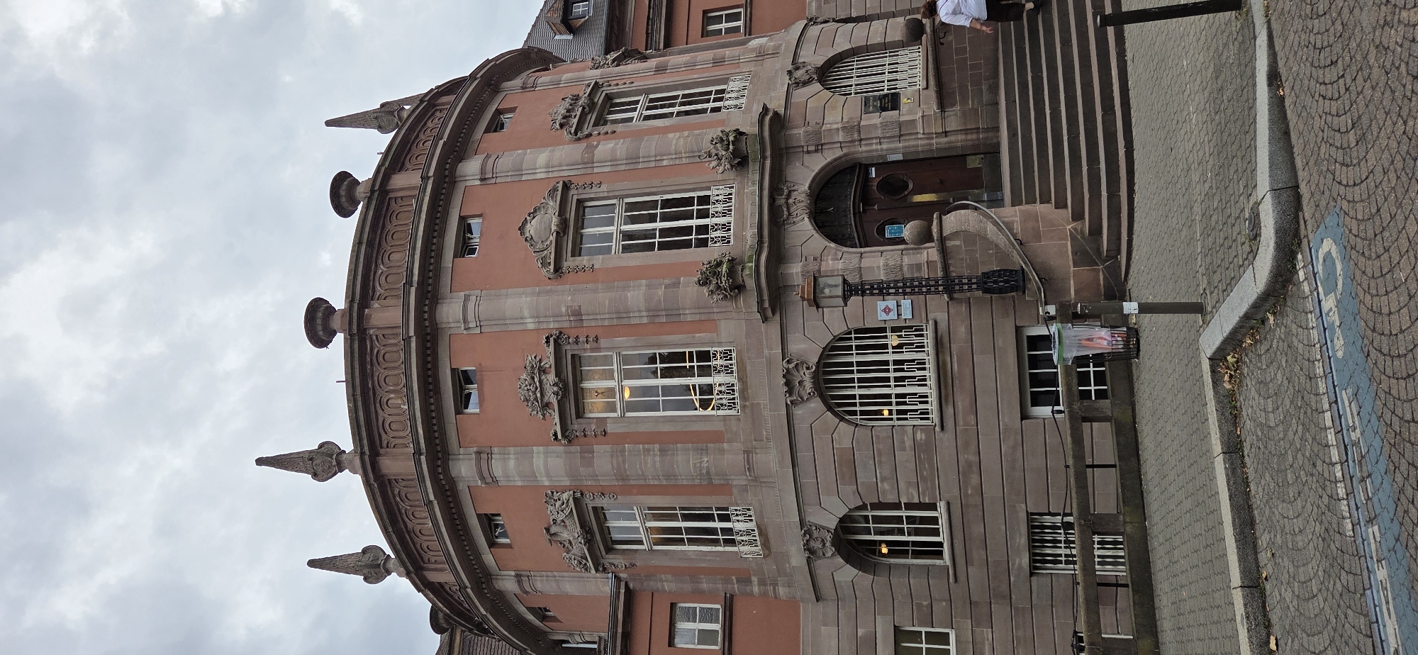 The image shows the facade of a large, circular, pink-colored building with ornate stone accents. The building has multiple stories with arched and rectangular windows, many of which are barred. Decorative carvings are present above some of the windows. A large wooden door is located at the base of the building, accessed by a wide set of stone stairs. A lamppost stands to the left of the entrance. In the foreground, a paved area with cobblestones and a disabled parking symbol is visible. A person in a white shirt and brown pants is walking up the stairs on the right side of the image. The sky is overcast. This building is identified as the Palace of Justice in Strasbourg, France.