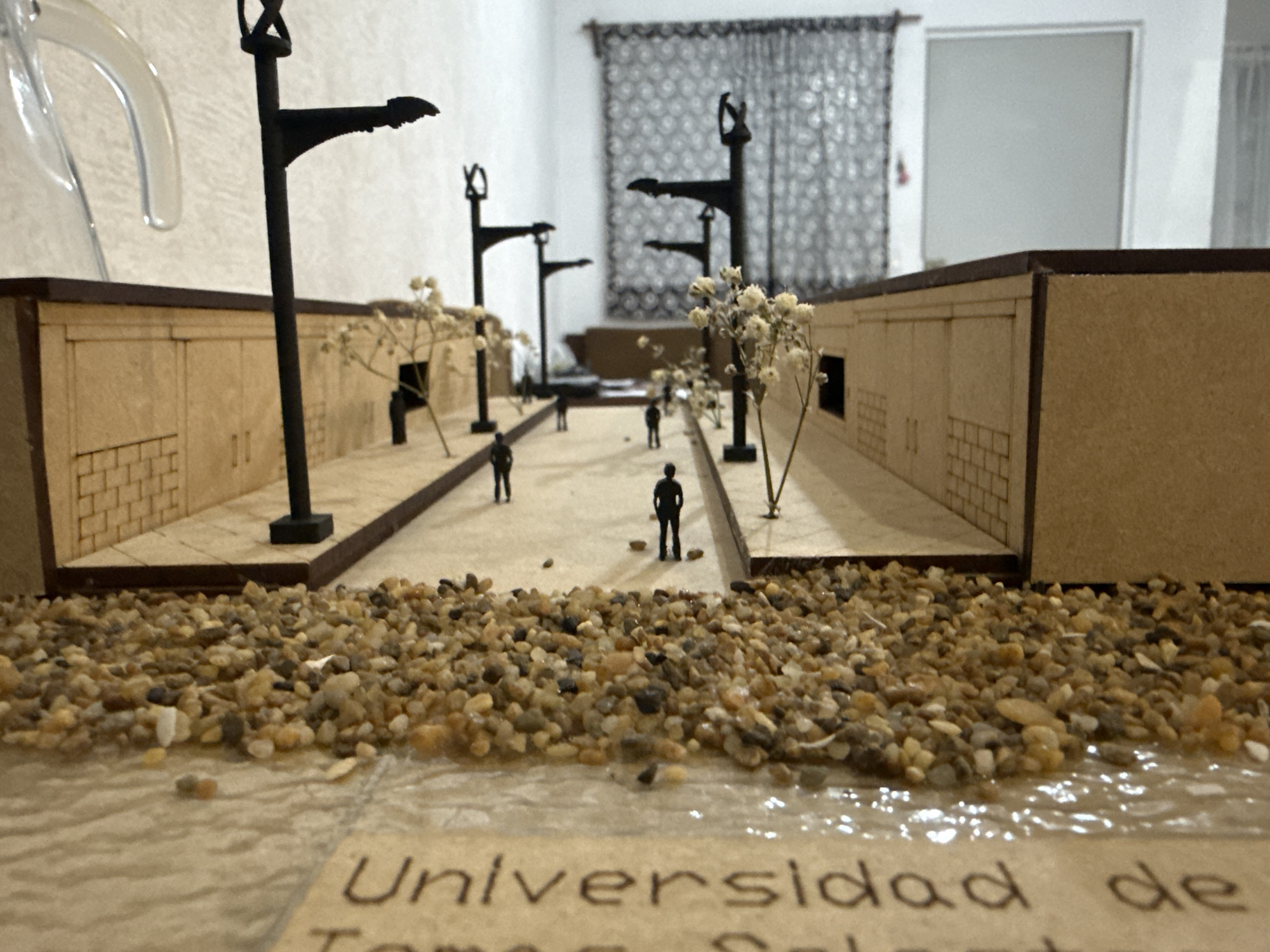 This medium shot captures a detailed miniature architectural model, likely a diorama, depicting a street scene. The model features two rows of light brown, wooden buildings with carved details resembling brickwork, doors, and windows, lining a central street. Black miniature streetlights are strategically placed along the street and sidewalks, some with small, dried white flowers or foliage clustered at their bases, representing trees or bushes. Several dark silhouette figures, representing people, are scattered along the street and sidewalks, adding a sense of scale and human presence to the model.
In the immediate foreground, a textured surface resembles a shallow body of water with small, natural-colored pebbles and stones forming a bank or shoreline. The water surface appears glossy and slightly rippled. Partially visible text is inscribed on a lighter surface beneath the water feature, clearly showing "Universidad de" followed by partially visible words like "Tomo Salat" or similar, suggesting an academic project or display related to a university, possibly in Puebla, Mexico.
The background reveals an indoor setting with a light-colored wall, a dark patterned curtain, and what appears to be a window or doorway. A clear, curved plastic object is visible in the upper left background, unrelated to the model itself. The lighting is even, suggesting an indoor environment. The perspective emphasizes the miniature world, with the foreground elements close to the viewer and the street receding into the distance, creating depth.