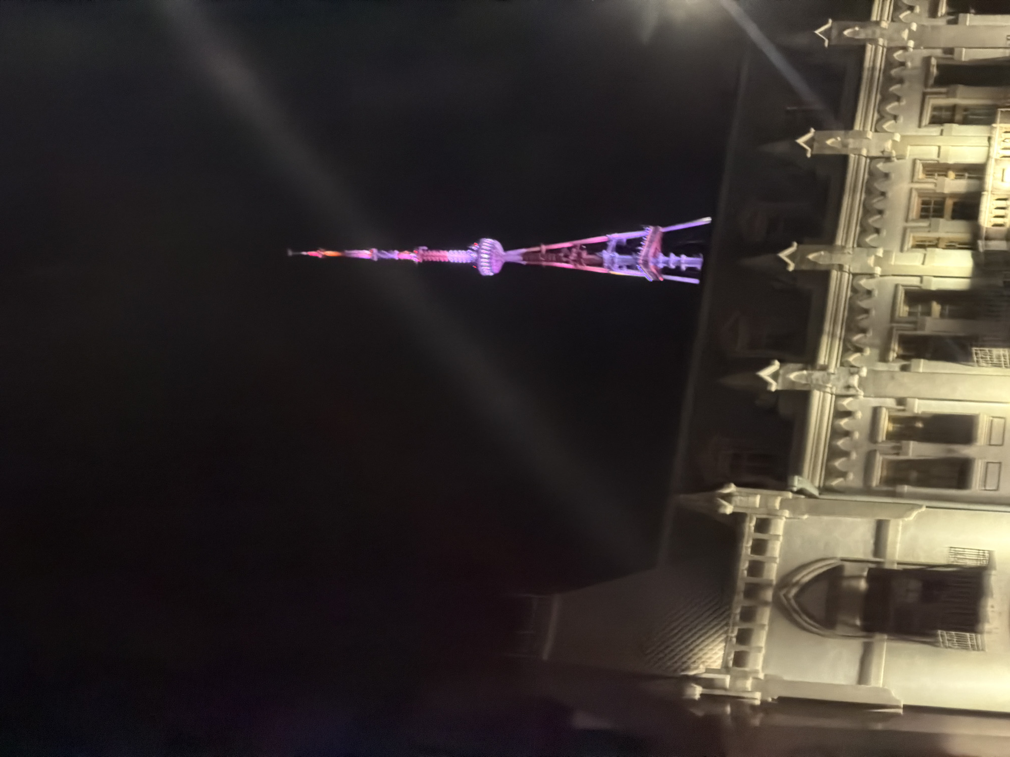 The image shows the Tbilisi TV Broadcasting Tower in Tbilisi, Georgia at night. The tower is illuminated with pink and purple lights. Part of a building with arched windows and decorative ledges is also visible. The sky is dark.