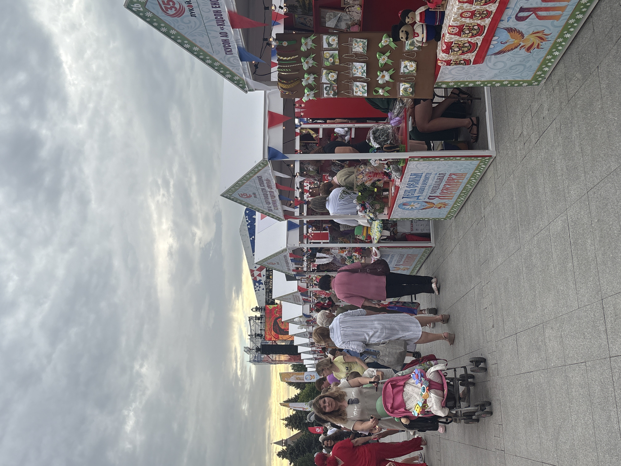 An outdoor market or fair is depicted under an overcast sky. A crowd of people, including adults and a child in a stroller, are walking along a paved pathway. Stalls are set up on either side of the pathway, selling various goods such as crafts, jewelry, and dolls. Some stalls have festive decorations like pennant banners. In the background, a large stage with a screen is visible, suggesting a performance or event is taking place. The overall scene conveys a lively atmosphere of a public gathering. The text on the stalls and banners appears to be in Cyrillic script, indicating a Russian location. The mention of "Astrakhan, Russia" in the prompt suggests this specific location.