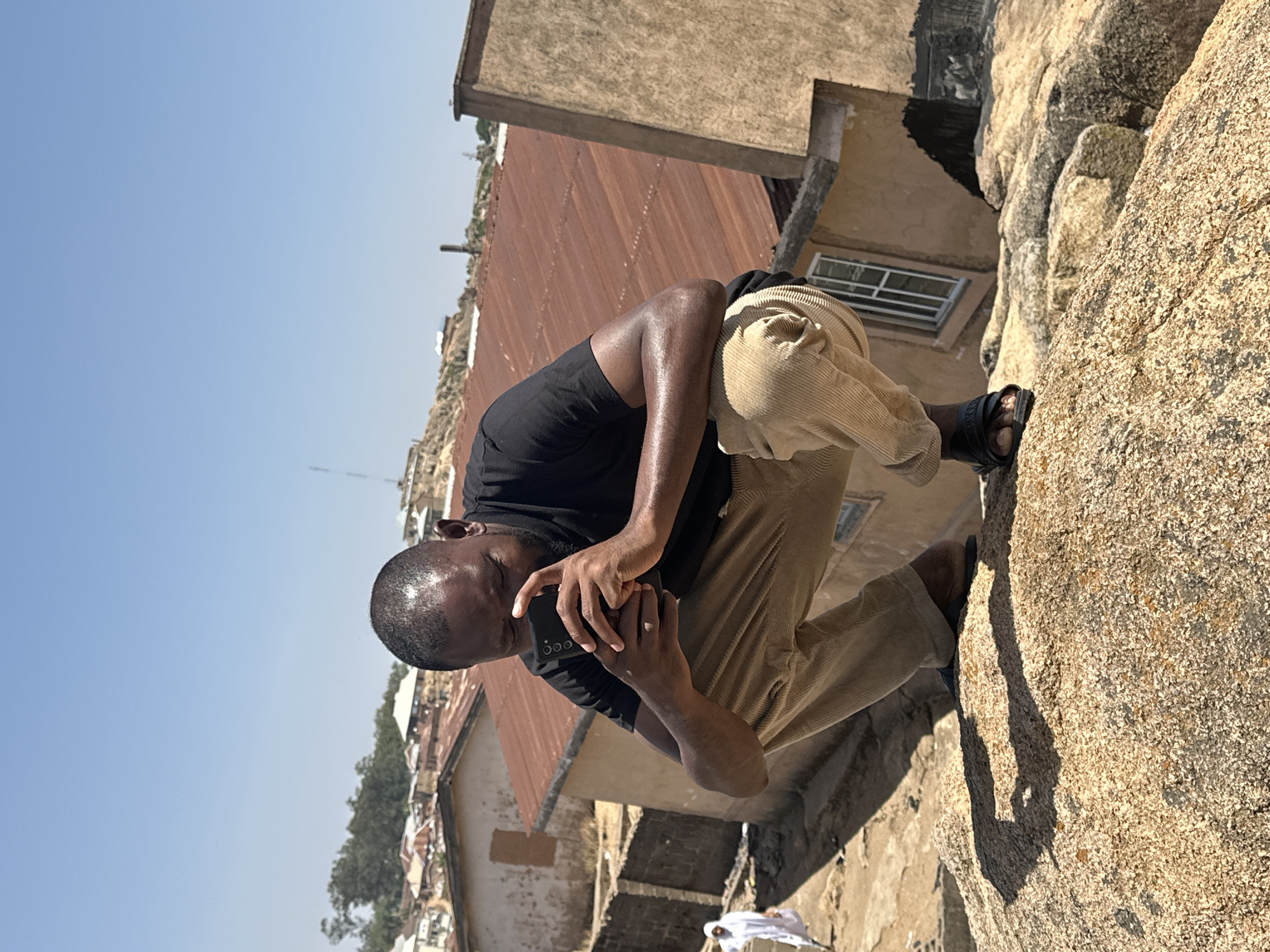The image captures a man outdoors in Jos, Nigeria. He is kneeling on a rock formation, holding a smartphone in his hands. He is wearing a black t-shirt, corduroy pants, and black sandals. The background shows a cityscape with buildings, rusty corrugated iron roofs, a window, and foliage visible in the distance under a clear sky. A white cloth lies on the ground near the rock formation.
