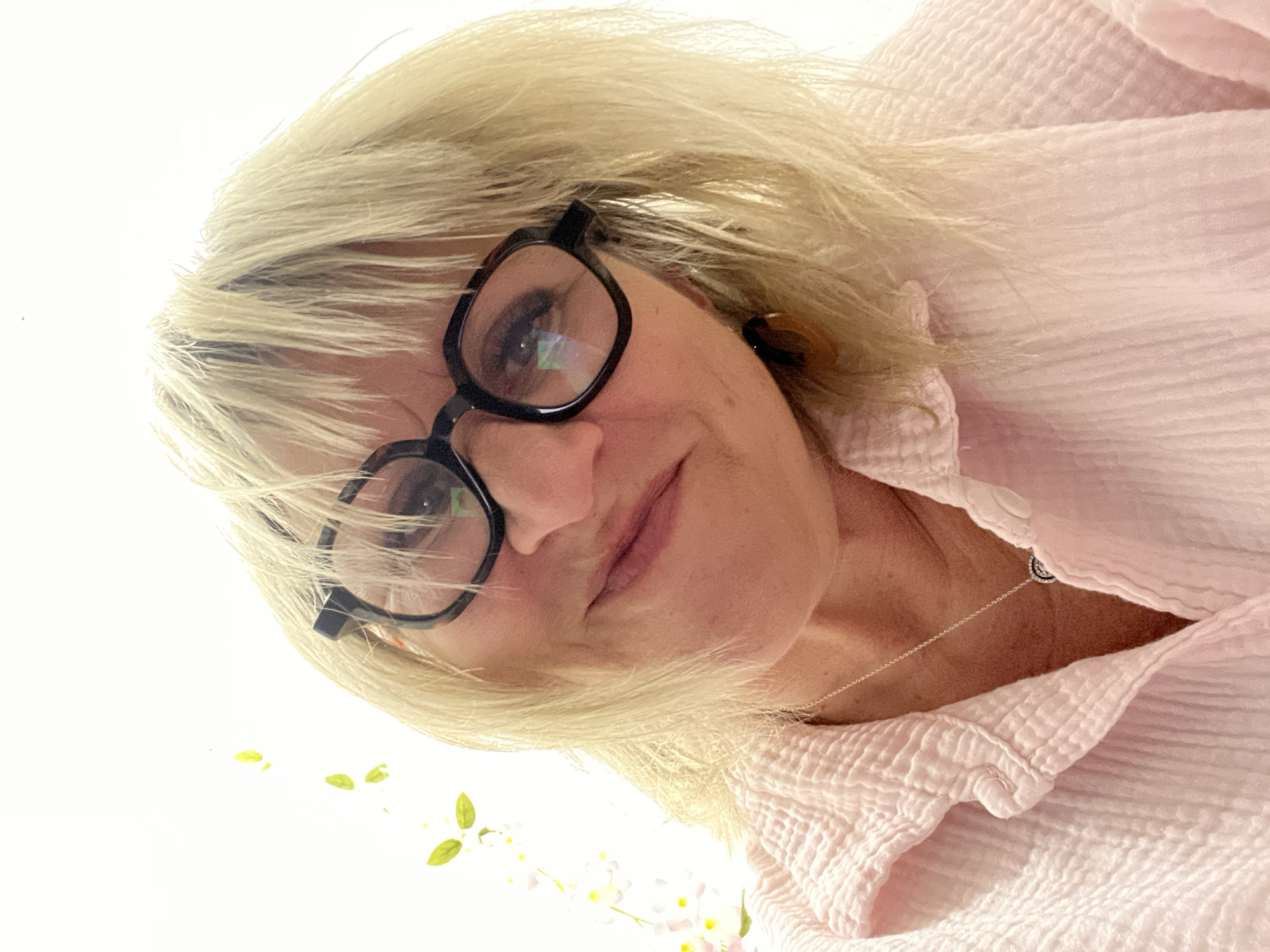 A woman with blonde hair and black-rimmed glasses is seen from a low-angle perspective. She wears a light pink, textured shirt and a silver necklace. The background is a bright white, with a few green leaves and white flowers visible in the lower portion of the frame. There is no information available in the image that identifies the city of Montélimar, France.