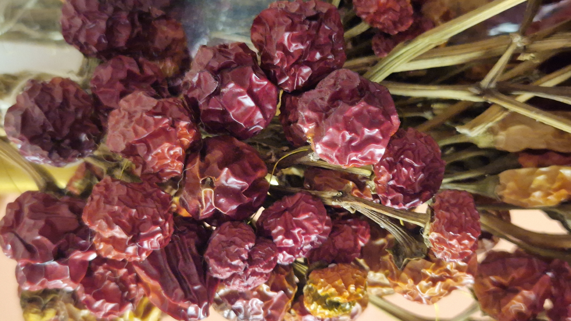 The image shows a close-up view of a bunch of dried, red chili peppers. The peppers are small and round, with wrinkled skins, and are attached to thin, dried stems. Some of the peppers are a deep red, while others are a lighter, more orange-red hue. The background is softly blurred, with warm, peachy tones. The composition is tight, filling the frame with the texture and color of the peppers. No specific location in Taranto, Italy is identifiable from the image.