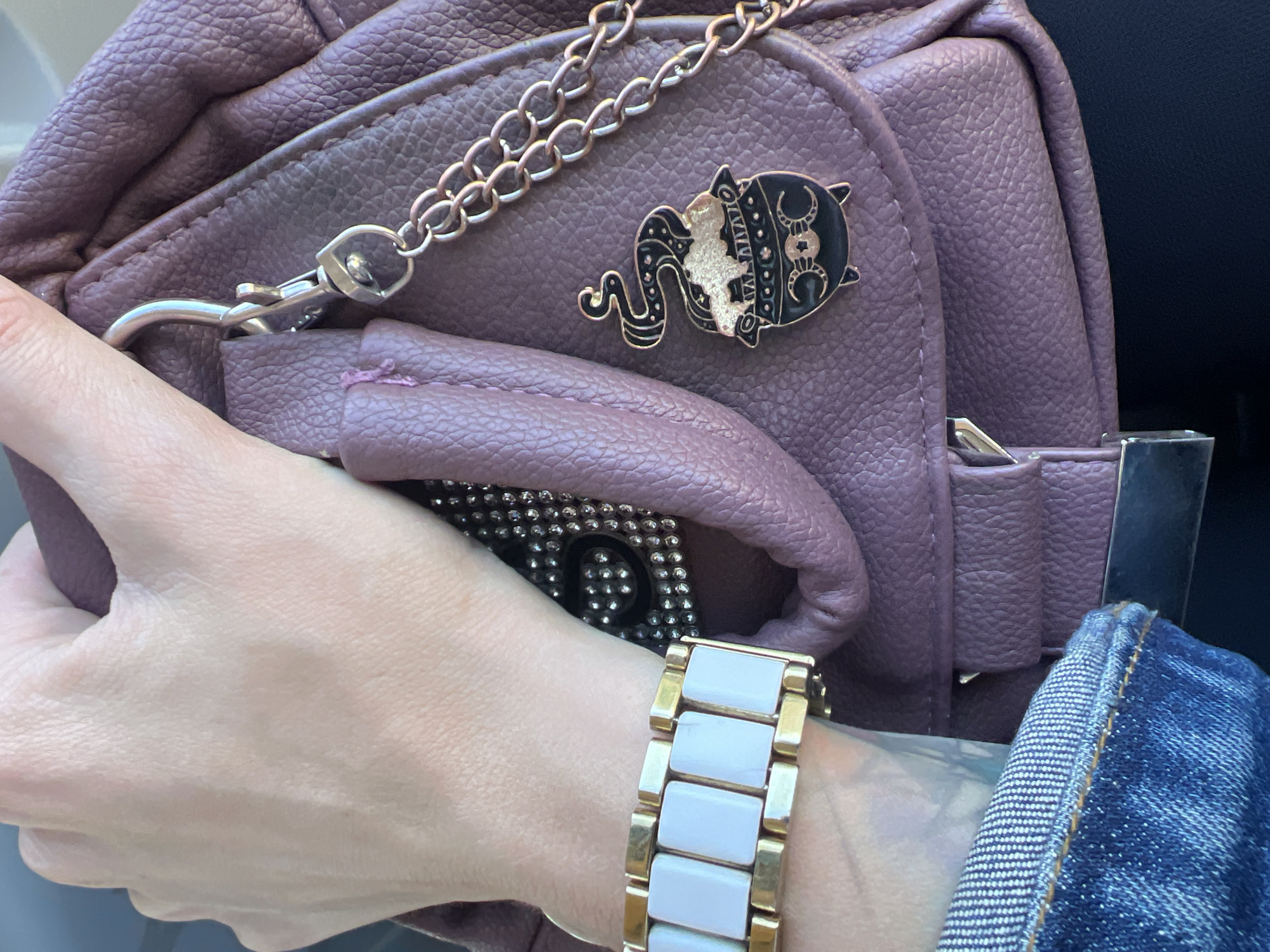 The image shows a close-up of a person's hand holding a purple leather purse. The purse has a chain attached to it and a charm of a cauldron with smoke. The person is wearing a bracelet with white rectangular links and a gold band. Part of a denim jacket is also visible. The scene does not contain any information about the location in the city of Krasnoe-na-Volge, Russia.