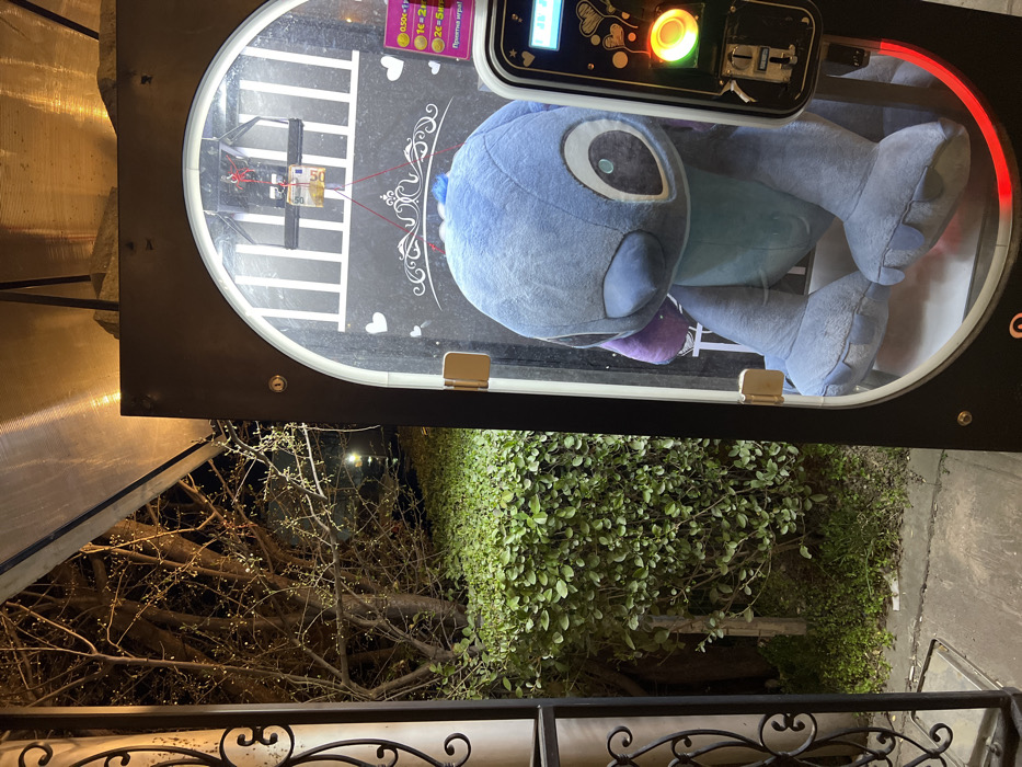 A nighttime outdoor scene depicts a black claw game machine positioned to the right of the frame's center. The machine features a large, oval-shaped clear viewing panel revealing a substantial, light-blue plush toy resembling Disney's Stitch, seated within the lower compartment. Above the plush, a yellow 50 Euro banknote is suspended by thin red strings or wires from a transparent claw mechanism. The internal back panel is black with vertical white stripes and white heart-shaped decals. A control panel on the right side of the machine includes a coin slot, a yellow button, a green button, and a digital display. Text in Bulgarian on this panel details pricing ("0.50лв=1лв", "1лв=2лв", "2лв=5лв") and a greeting ("Приятна игра!").
To the left of the machine, a decorative black metal railing with scrollwork is present in the foreground. Beyond this, a dense, well-trimmed green hedge approximately one meter in height extends across the mid-ground. Behind the hedge, deciduous tree branches with small, light-colored buds are visible against the dark background, illuminated by distant light sources. The ground beneath the hedge appears as dark soil or sparse grass with minor debris. An overhead translucent corrugated plastic awning, illuminated with an orange glow, partially covers the top portion of the scene.
The presence of Bulgarian lev (лв) currency conversions on the claw machine confirms the location is in Bulgaria. The specific address or neighborhood within Sofia, Bulgaria, is not discernible from the visual information provided.