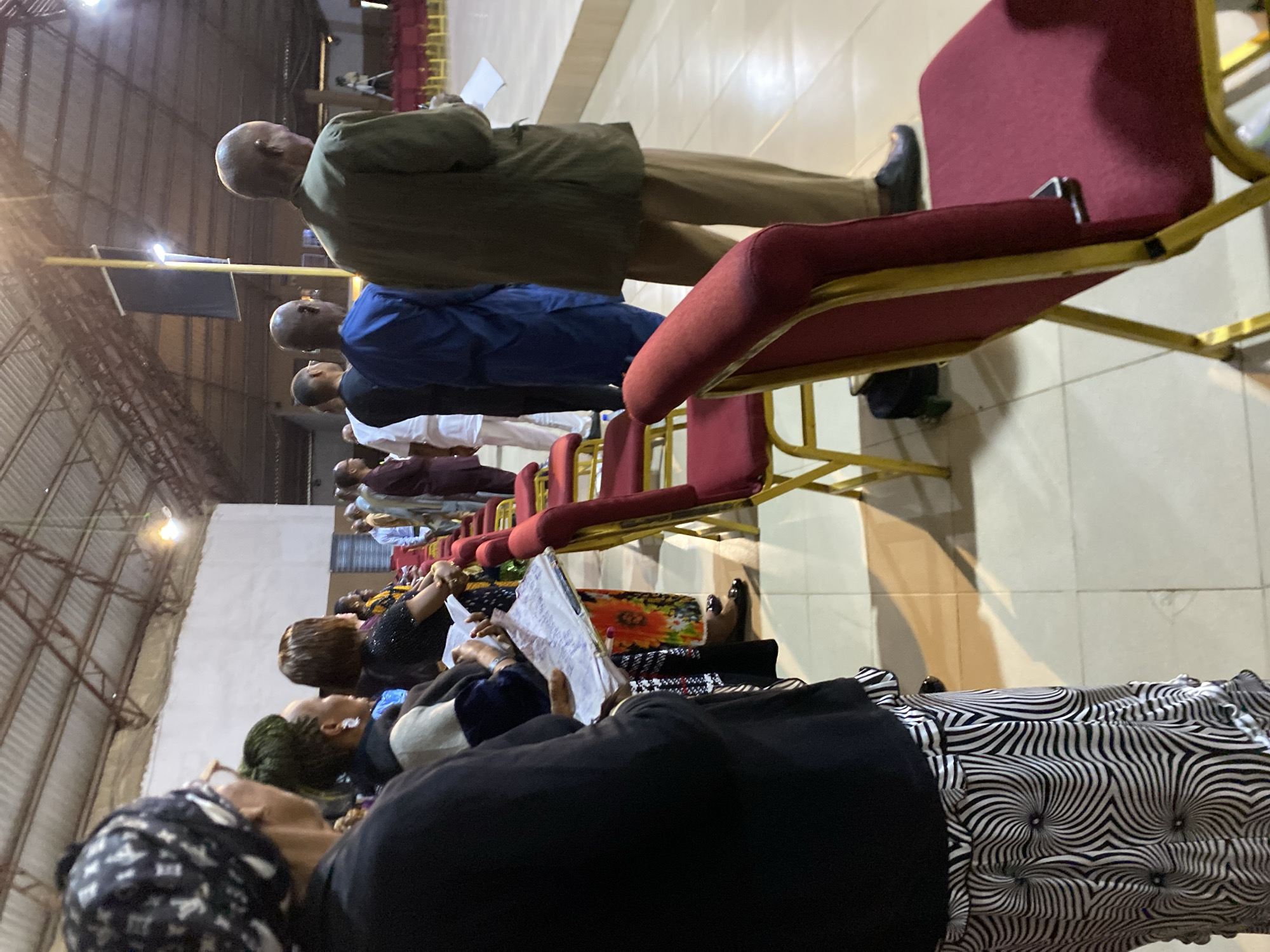 The image shows a group of people standing in a church hall on Rukuba Road, Nigeria. The hall has a high, white ceiling with exposed beams. Red chairs with gold frames are arranged in rows. Some people are holding papers and appear to be engaged in a service or ceremony. The lighting suggests it might be evening or indoors with artificial lights. The overall atmosphere seems solemn and respectful.