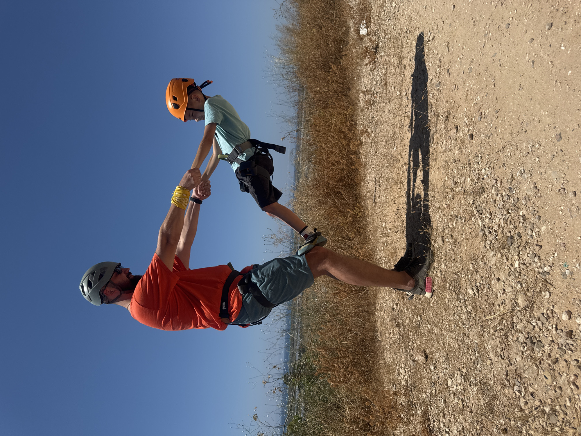 A father and son stand on a path in the Hof HaCarmel Regional Council, Israel. The father, wearing a gray helmet and an orange shirt, holds the son in the air by his hands. The son wears an orange helmet and a light blue shirt. Both are wearing climbing harnesses. The father is balancing on one foot on a grassy, dirt path next to a dry field. A shadow of the pair is cast on the path. The sky is clear and blue.