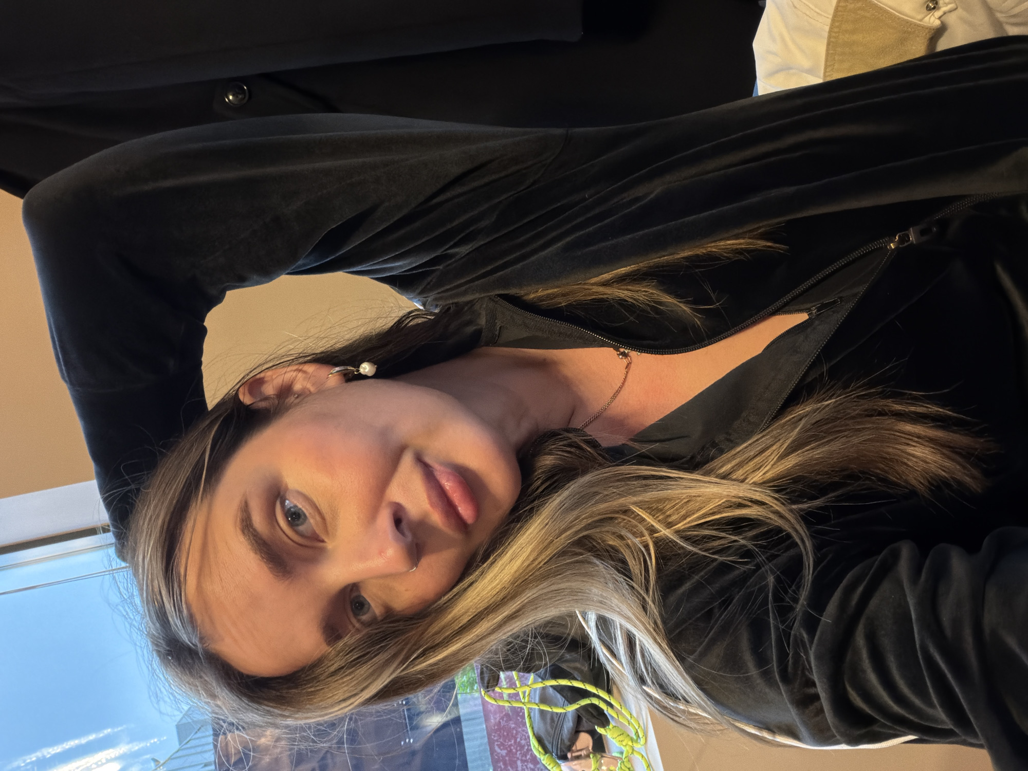 A woman with light blonde-streaked brown hair is posing for a selfie. She is wearing a black zippered jacket and has a nose ring and earrings. Her hair is flowing down her shoulder. The background shows an interior setting with a window, offering a view of a city. Based on the architecture visible through the window, and the style of the buildings, it is plausible that the location is in Moscow, Russia. However, without specific recognizable landmarks or context, it's difficult to confirm the exact location definitively.