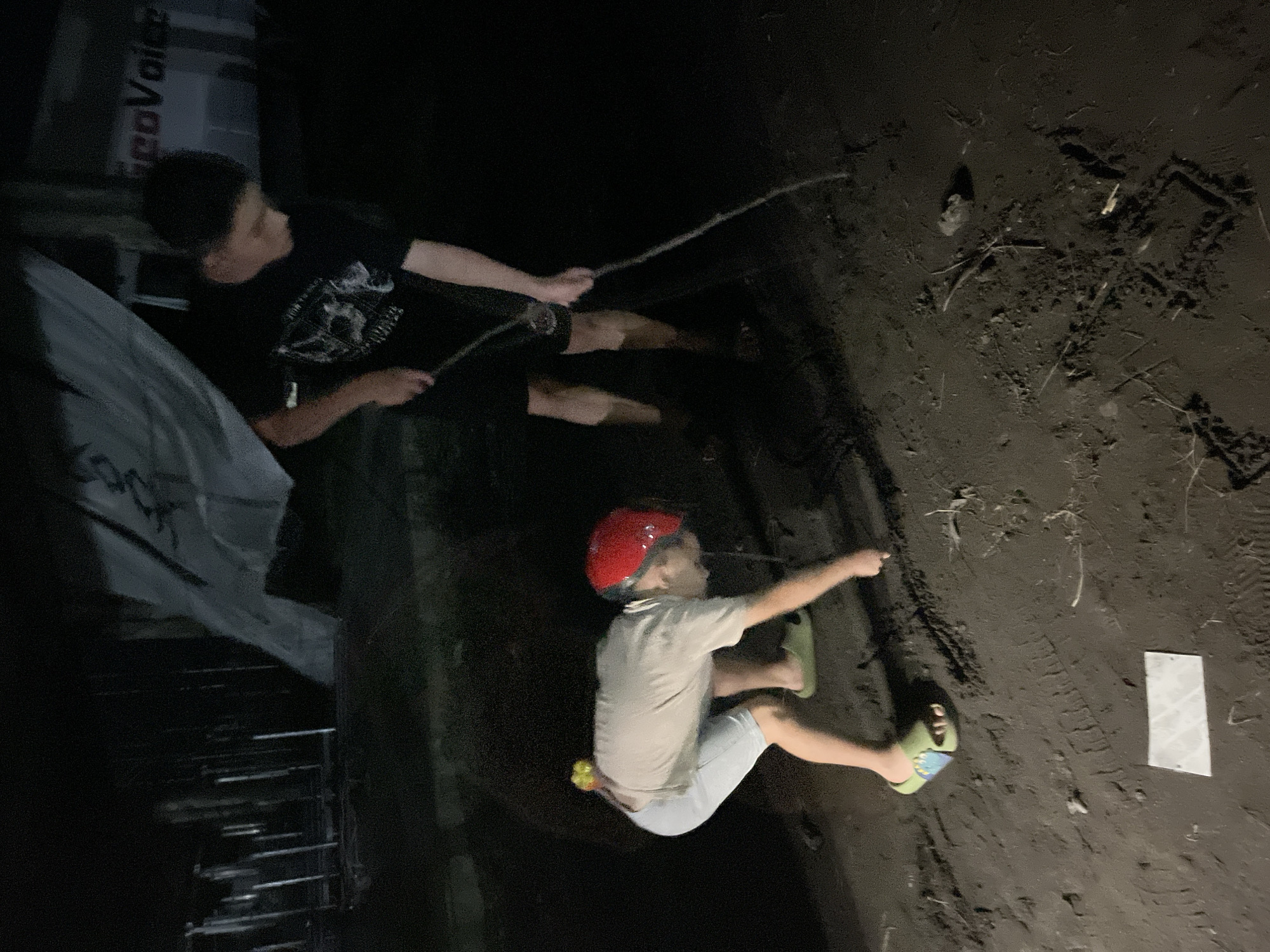 In Poti, Georgia, at night, two individuals are engaged in an activity on a dirt surface that appears to be an outdoor area. The person in the foreground is a child wearing a red helmet, a light-colored t-shirt, and light-colored shorts. They are bent over, seemingly interacting with the ground. Attached to the child's waistband are two yellow objects. They are also wearing green slip-on sandals. The second individual, in the background, is a youth wearing a black t-shirt with a graphic on it and dark shorts. This individual is holding a stick and appears to be drawing or marking the dirt. The overall environment is dark, with some ambient light illuminating the scene, creating shadows and obscuring details. The ground is covered in dirt and debris, with visible tire tracks or footprints in some areas. In the background, there is a structure with a white and grey banner or tarp, and signage with red lettering that is partially visible. The scene suggests an outdoor recreation or play setting at night.