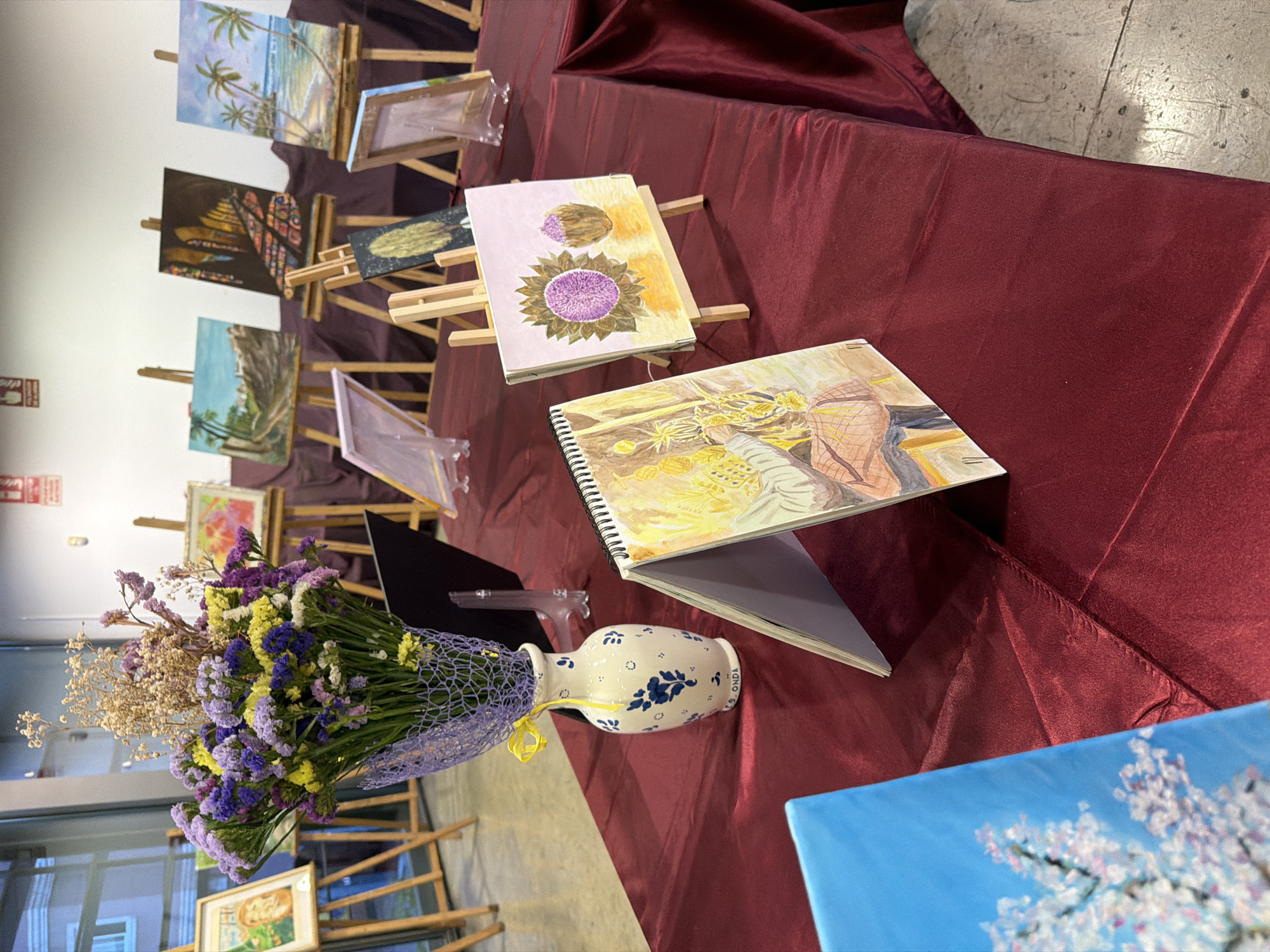 A table draped with a deep red cloth displays an art exhibition. In the foreground, a vibrant bouquet of purple, yellow, and white dried flowers is arranged in a blue and white patterned ceramic vase. To the right of the vase, an open sketchbook with a watercolor painting of a figure is propped up on an easel. Beside it, another painting on an easel features two purple thistle-like flowers against a yellow background. Further back, more paintings are displayed on wooden easels, including landscapes and a beach scene with palm trees. A blue canvas with a painting of white blossoms is partially visible in the lower left corner. The background suggests an indoor venue with white walls and a window. The overall scene depicts a small art display.