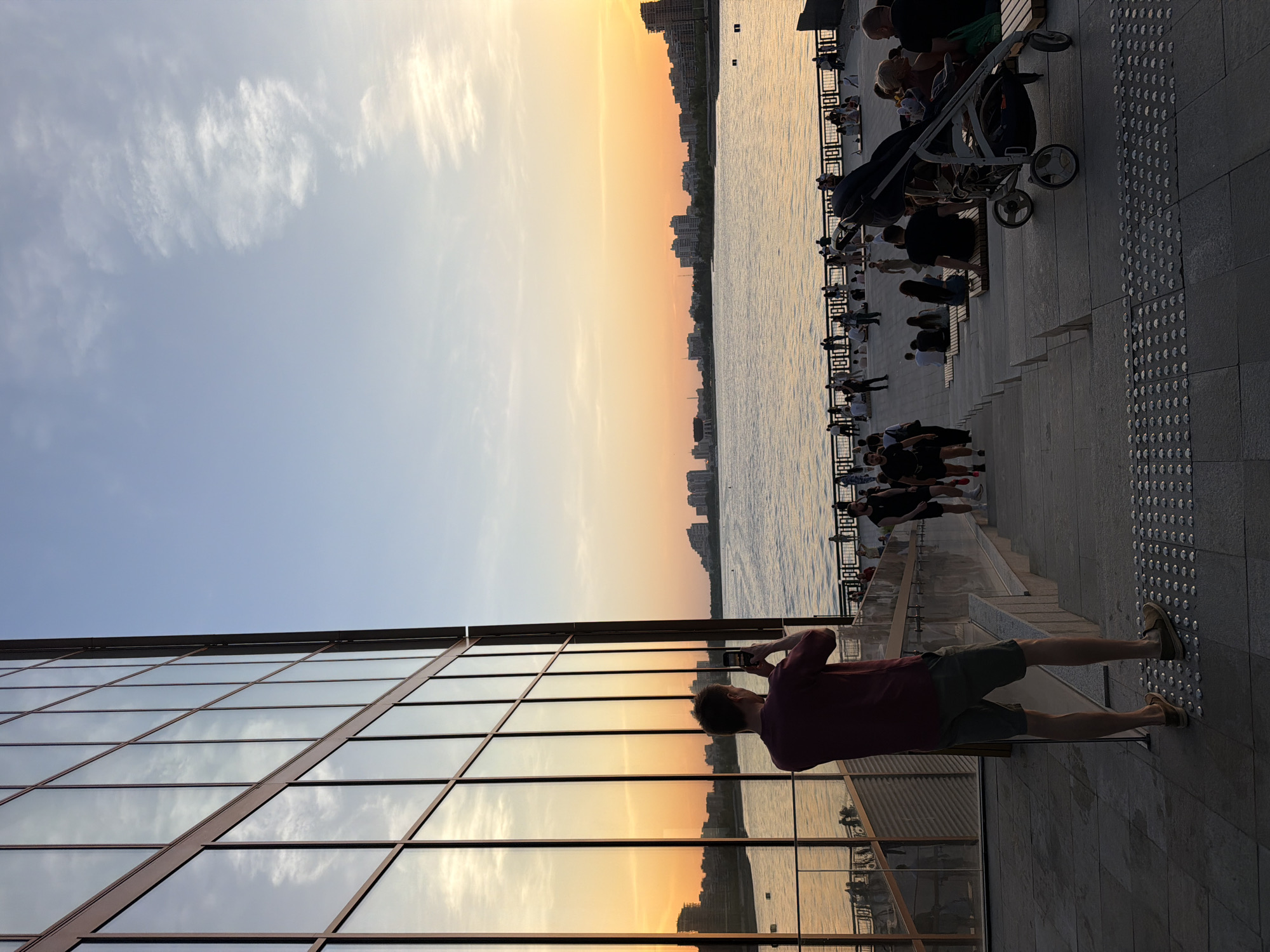 The image captures a scene along a waterfront in Kazan, Russia. The sky is bright with a sunset, casting a warm glow on the water and reflecting in the glass windows of a building in the foreground. On a paved area adjacent to the water, people are relaxing and socializing on benches and steps that lead down to the water's edge. A stroller is parked nearby, suggesting families are present. A man stands near the building, facing the water and taking a photo with his phone. The waterfront features a metal railing separating the seating area from the water. The background includes the cityscape across the water.