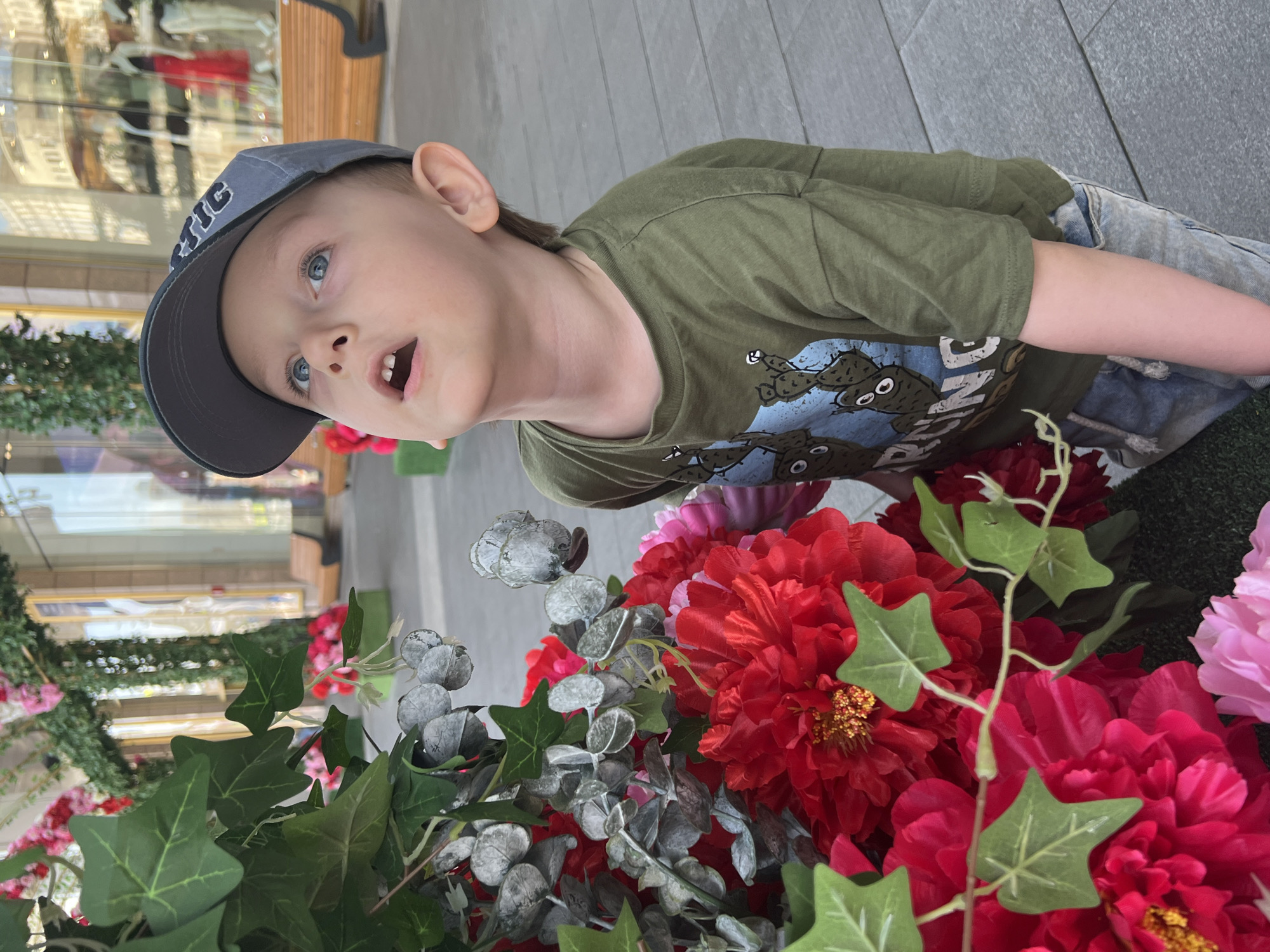 A young male child, estimated to be between 3 and 5 years old, is depicted in a kneeling or crouching position, looking sharply upwards with his mouth slightly agape, indicating an expression of curiosity or surprise. He has fair skin, blue eyes, and light brown hair visible beneath a grey baseball cap with white lettering that partially reads "ARCTIC". He is wearing an olive-green t-shirt with a light blue graphic featuring cartoon cacti and the word "RUNNING", paired with light blue denim shorts.
The child is positioned next to a large, dense display of artificial flowers and foliage. This arrangement includes numerous large, deep red peony-like blossoms, vibrant pink flowers, and various artificial green leaves, specifically dark green ivy-like leaves and silvery-grey eucalyptus-like leaves with spherical seed pods. A section of artificial green turf is visible at the base of this floral display.
The background reveals a modern pedestrian shopping street, paved with grey rectangular tiles. The street is lined with multi-story commercial buildings featuring large glass display windows that reflect the bright sky, architectural details, and interior retail displays including mannequins, one prominently dressed in a red gown. More artificial greenery, resembling garlands, is draped across structures above the shop windows, and additional pink artificial flowers are visible further down the street. Several light-colored wooden benches with dark metal frames are spaced along the walkway. The scene is situated in a commercial or pedestrian area within Moscow, Russia.
