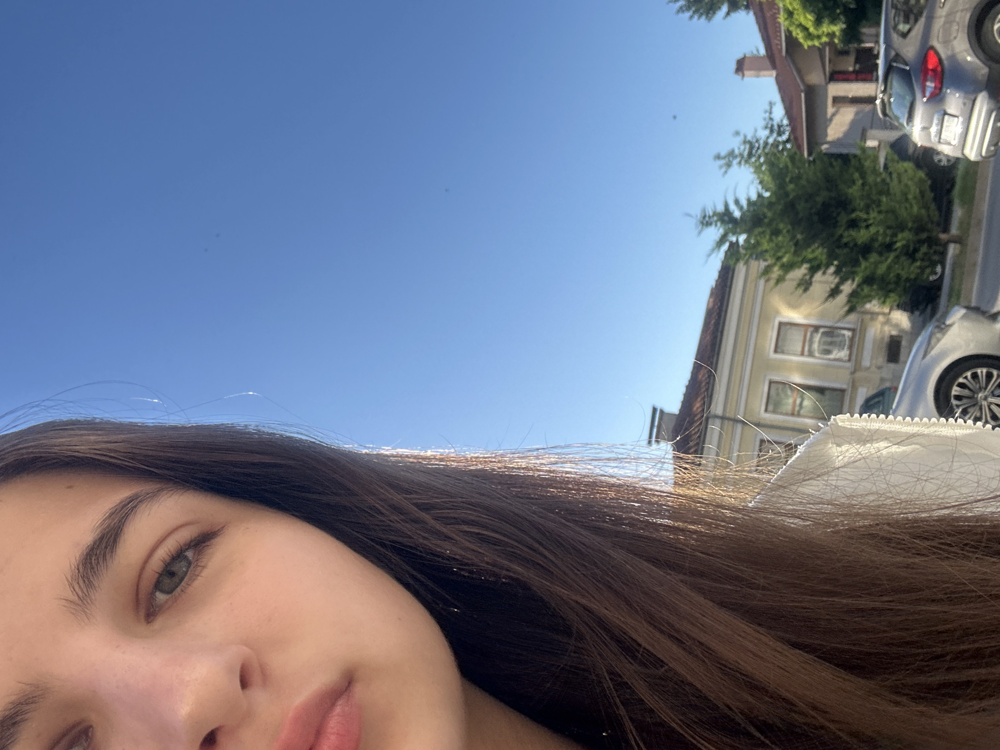 A close-up selfie shows a young woman's face and shoulder against a bright blue sky. Her left eye, nose, and lips are visible, with her long brown hair cascading down her shoulder. Sunlight glints off her hair. In the background, a street scene unfolds with houses, trees, and parked cars. The architecture suggests a European setting. The location is identified as Shumen, Bulgaria.