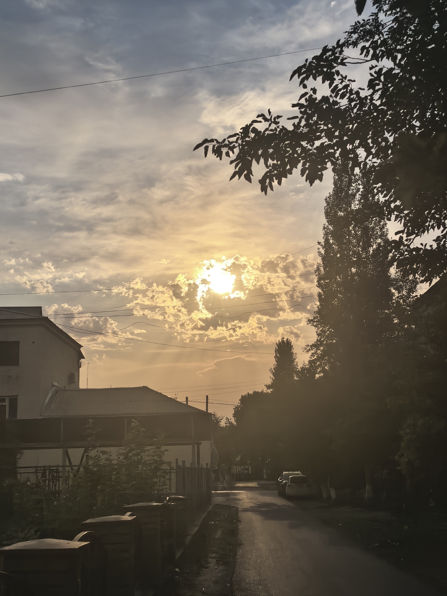 The image depicts a residential street scene in Fergana City, Uzbekistan, during sunset. The sky is filled with a mix of gray and white clouds illuminated by the setting sun, which is partially obscured by clouds. The sun's rays create a golden hue across the sky and cast long shadows. On the left side of the street, a two-story building with a tiled roof is visible, along with a smaller structure with a porch. A fence and some vegetation separate the property from the street. The street itself is paved and appears wet, possibly from recent rain. Several trees line the right side of the street, their foliage silhouetted against the bright sky. Two cars are parked on the side of the street, partially visible through the trees. The overall atmosphere is tranquil, with the warm colors of the sunset creating a serene mood.