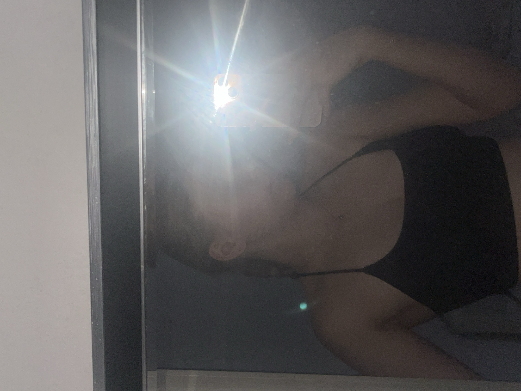A person is visible in a mirror reflection, holding a smartphone to take a selfie. The person is wearing a black tank top. The background appears to be an indoor setting, possibly a room, with a light source causing a bright glare in the reflection. The image does not provide any specific details that would allow for the identification of the city of Perm, Russia.