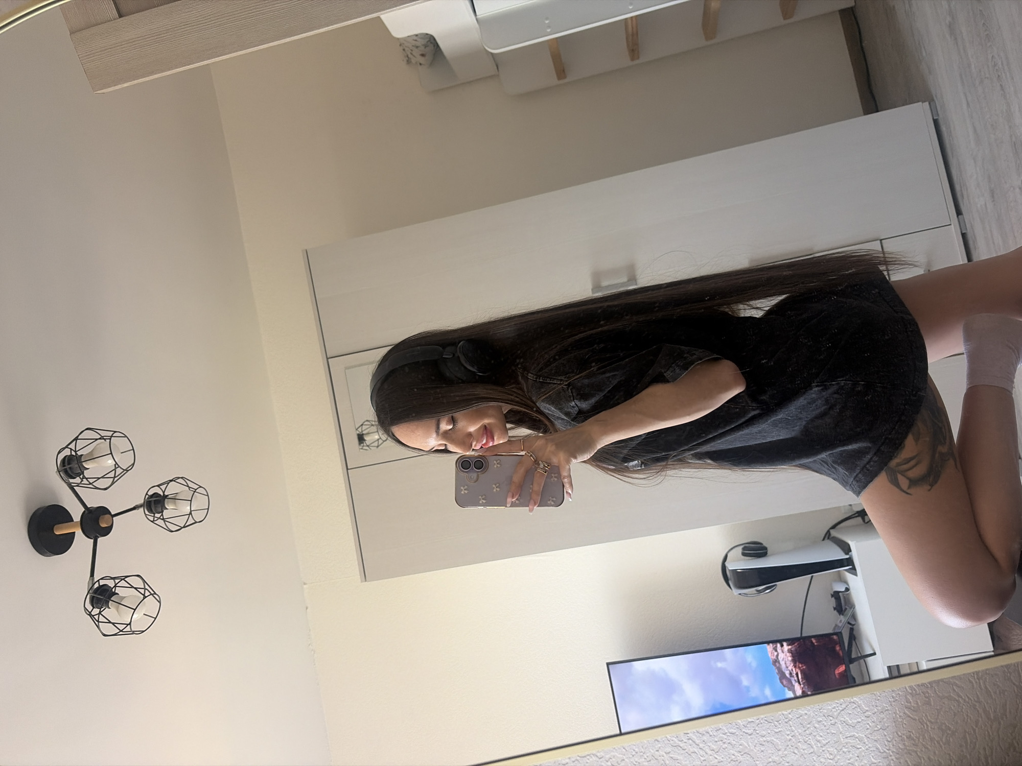 A young woman with long, dark hair poses in front of a mirror, taking a selfie with her mobile phone. She is wearing headphones and a black shirt. The woman is partially visible from the waist up; her legs are shown from behind and are bent at the knees. She has a tattoo on her left leg. The mirror reflects a room interior with white walls, a modern-style ceiling light fixture, a white cabinet with drawers, and a television displaying a landscape scene. A white gaming console is visible near the bottom right of the frame. There is no indication of location in Saint Petersburg, Russia.