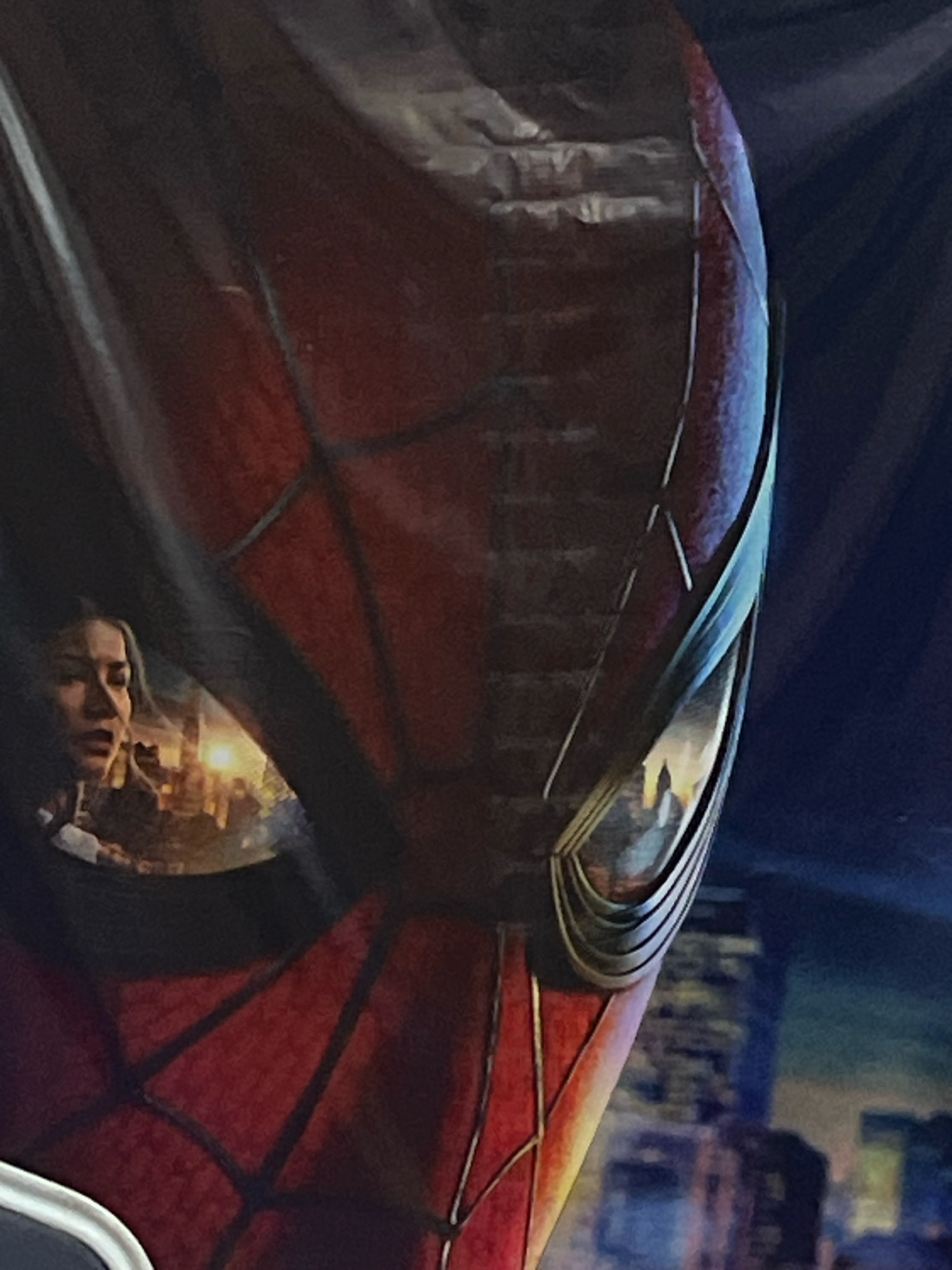The image is a close-up shot of a portion of the Spiderman costume, specifically the mask and part of the torso. The mask is primarily red with black webbing. In the left lens of the mask, a reflection of a woman's face is visible. She appears to be looking to her left with a surprised or concerned expression. Behind her, a blurry cityscape with illuminated buildings is visible. The overall scene appears to be a promotional image or a depiction of Spiderman with a city backdrop. The image does not provide any specific details about the city of Jos, Nigeria.