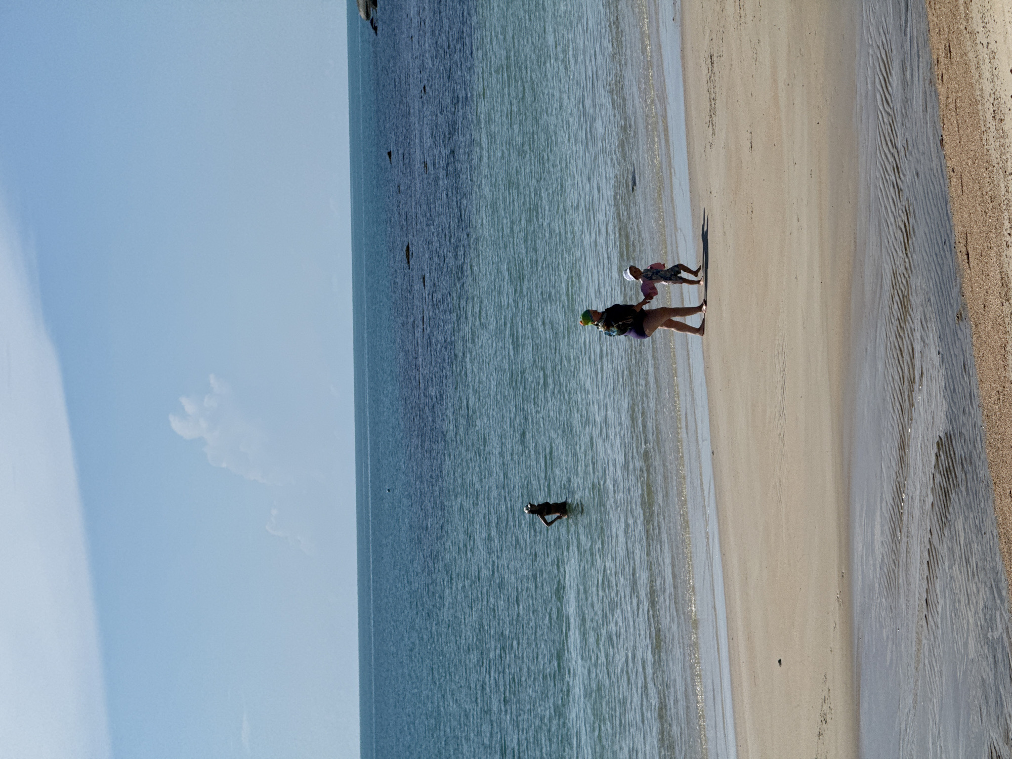 Two people, an adult and a child, walk hand-in-hand on a sandy beach. The adult, wearing a purple bathing suit and a colorful hat, leads the child, who wears a white hat and a patterned bathing suit with pink arm floaties. They are walking towards the right side of the frame, their shadows cast on the wet sand. In the shallow water to the left, a lone figure stands with hands on hips, partially submerged. The ocean stretches out to the horizon under a clear blue sky with a few wispy clouds. The beach is light brown sand with some ripples of water near the shoreline. The image depicts a scene in Baan Chaweng Noi, Thailand.