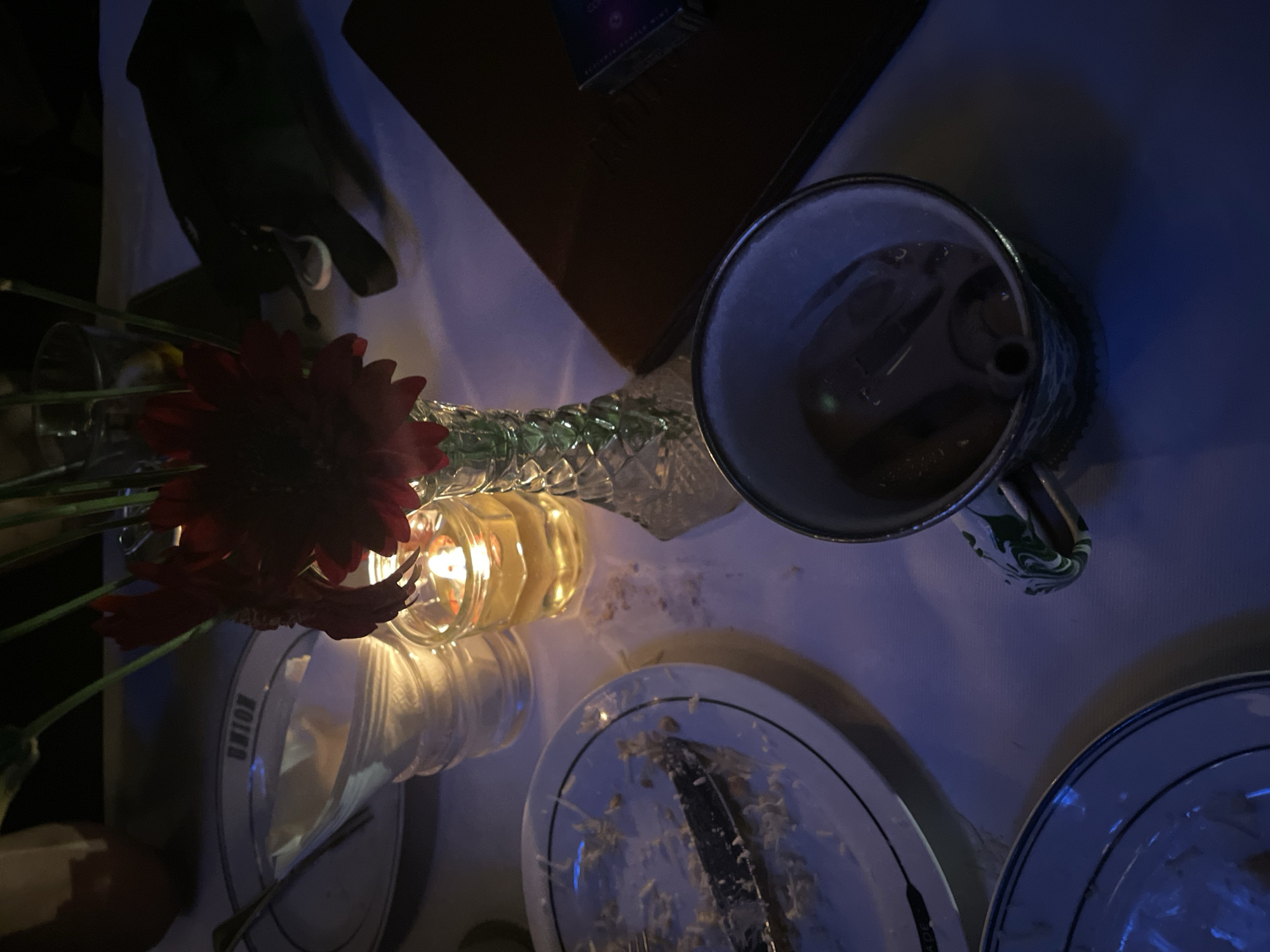 The image depicts an overhead shot of a dinner table setting. The table is covered with a white tablecloth. A red gerbera daisy and green foliage lie next to a crystal vase with two candle holders. One of the candle holders is lit. There is a mug with a green and white handle. At least two plates are visible, covered with food remnants. A knife is on one of the plates. There is also a brown book. The location is Central Jakarta, Indonesia.