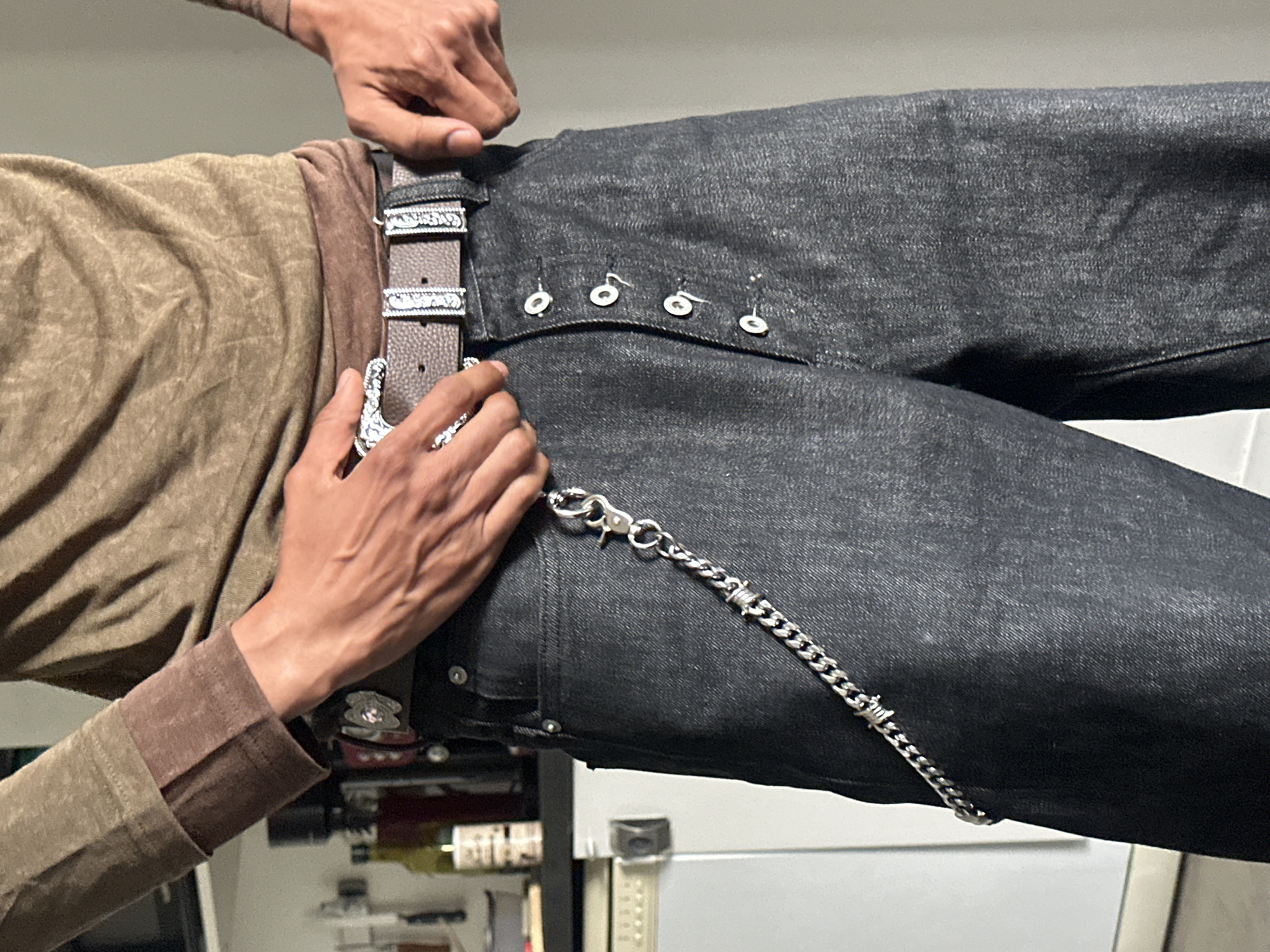 A man is adjusting a brown leather belt with ornate silver buckles around a pair of dark-wash denim jeans. The jeans feature a button fly and a silver chain attached to the front belt loop. The man is wearing a brown long-sleeved shirt. The background is cluttered with various items, including what appears to be a refrigerator and bottles, suggesting an indoor setting, possibly a home or apartment. The image does not depict Paris, France.