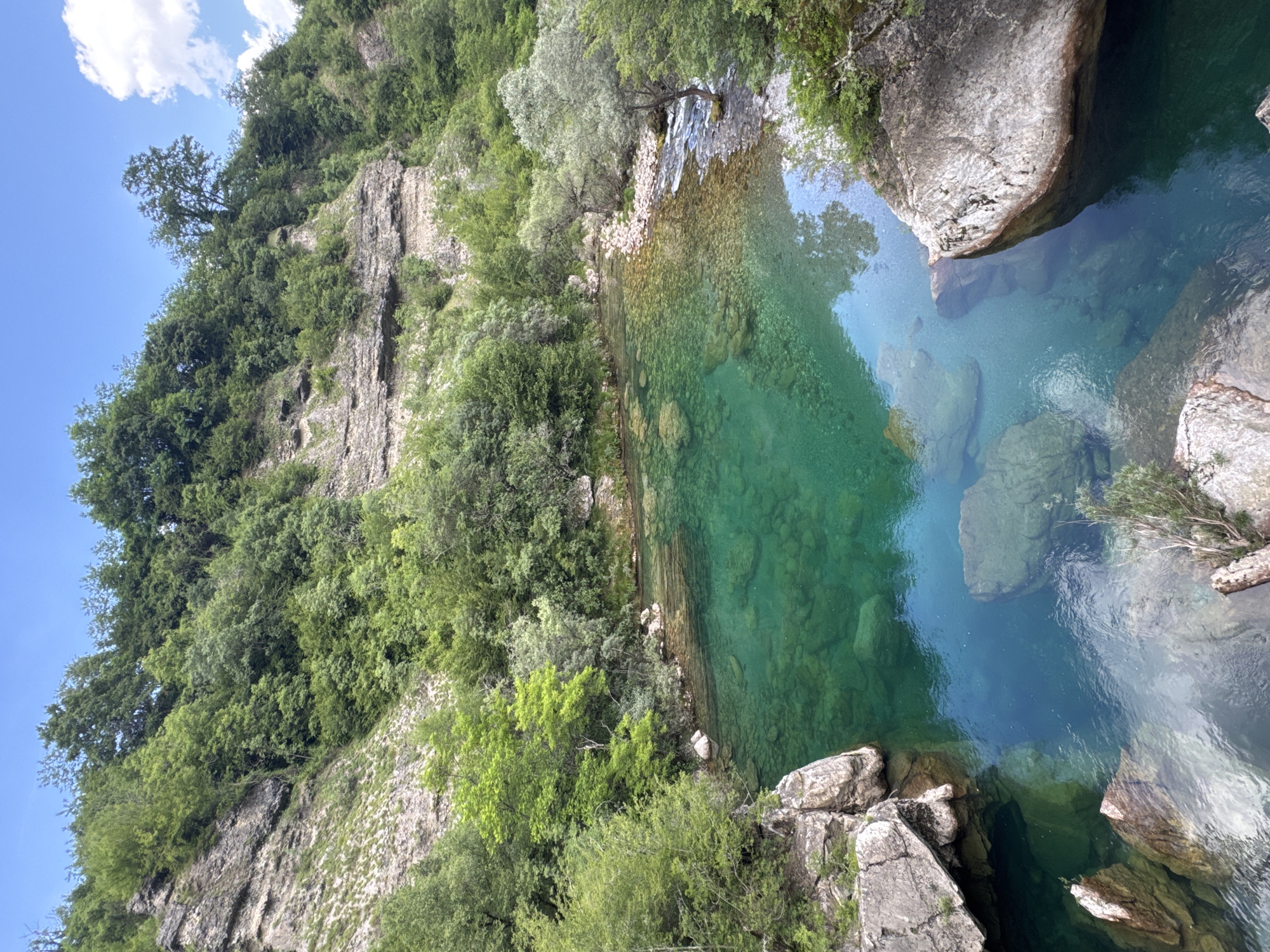 The image showcases a clear turquoise river in Međuriječje, Montenegro, surrounded by rocky cliffs and lush green vegetation. The water is so transparent that the rocks at the bottom are visible. The river is bordered by cliffs on both sides, heavily vegetated with trees and bushes. A mix of sunlight and shadows falls on the water and surrounding landscape.