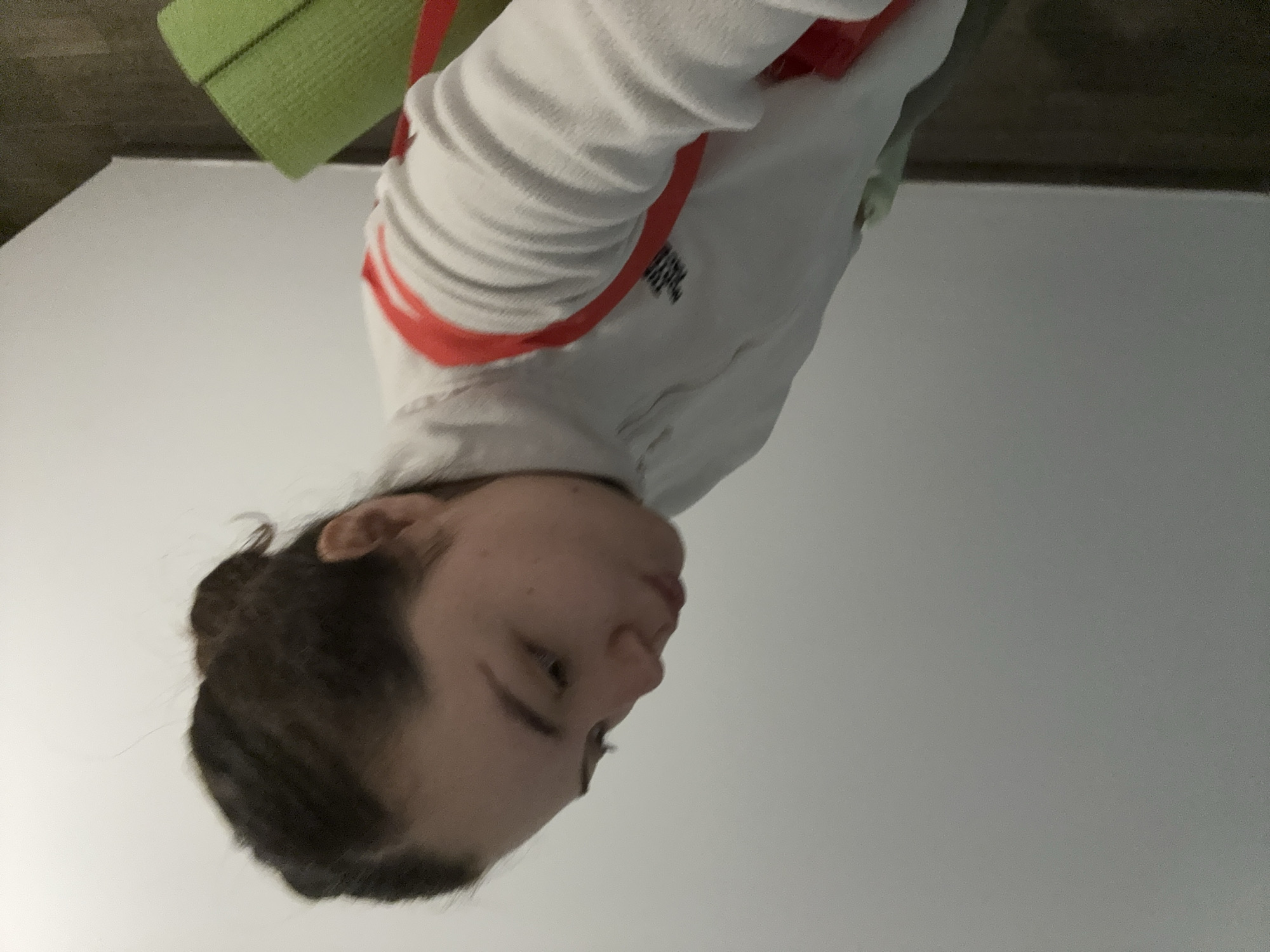 The image is an upside-down, close-up shot of a fair-skinned person with dark hair pulled back. They are wearing a white jacket with red stripes on the sleeves. A green object is visible behind the person's head. The background is a plain, light grey wall. There is no information to indicate that the image was taken in Mexico City, Mexico.