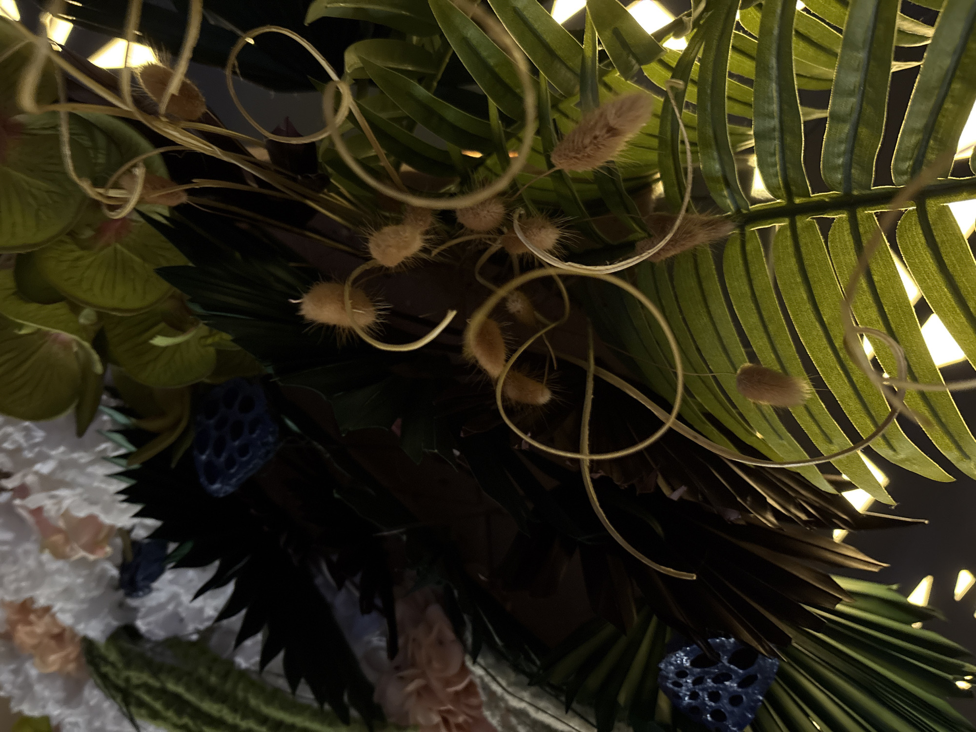 The image displays a close-up view of a decorative floral arrangement. The arrangement features a variety of artificial plants and flowers, including large, bright green palm leaves, darker, spiky leaves, and clusters of soft, fuzzy, oval-shaped elements that appear to be seed pods or dried flowers. There are also some delicate, light-colored flowers, possibly artificial carnations or hydrangeas, in the upper left portion of the image. A few dark blue, dried lotus seed pods are also visible. Thin, swirling tendrils or decorative wire elements are interwoven throughout the arrangement. The lighting suggests it is indoors, with warm, yellow lights visible behind the foliage, creating a soft glow and highlighting the textures of the leaves and pods. There are no people or animals present in the image. The location is not specified within the image itself and cannot be determined to be in Pleven, Bulgaria, or any other specific city.