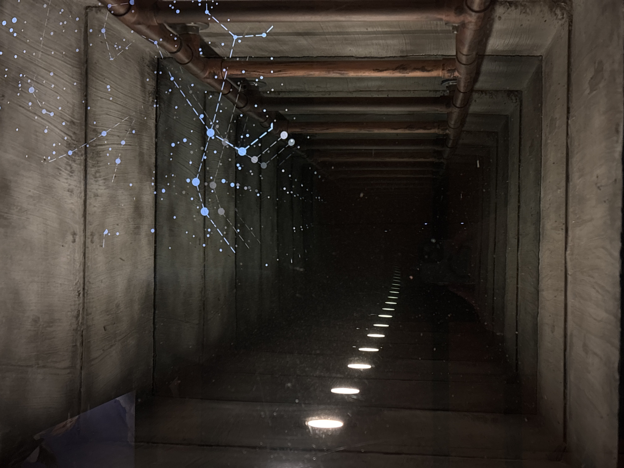 The image depicts a long, dark concrete tunnel with pipes running along the ceiling. A series of lights illuminate the floor, creating a linear perspective into the distance. An overlay of interconnected dots and lines is superimposed on the left side of the image, partially obscuring the view of the tunnel wall and ceiling. Based on the location and visual features, it is possible the image depicts an aqueduct. The presence of pipes suggests this might be a modern adaptation or modification of an older structure. It is unknown if any people are present within the tunnel.