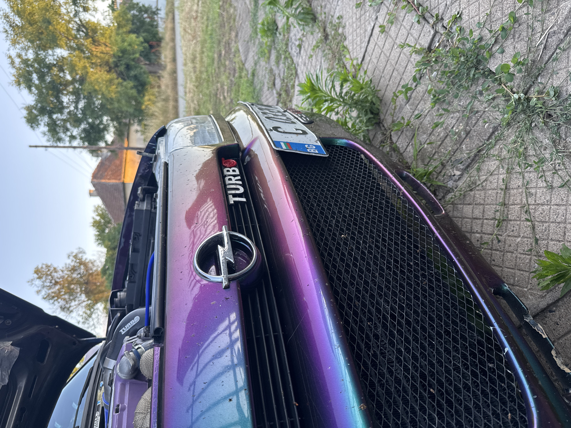 The image shows the front of a modified car with a vibrant purple and teal iridescent paint job. The hood is open, revealing engine components and blue hoses. A chrome Opel emblem is visible on the grille, and a "TURBO" badge is also present. A Bulgarian license plate, reading "C 0380 BG", is attached to the front bumper. The car is parked on a cracked concrete surface overgrown with weeds, in what appears to be a residential area with houses and trees in the background. The lighting suggests it is late afternoon or early evening. The location is identified as Lepitsa, Bulgaria.