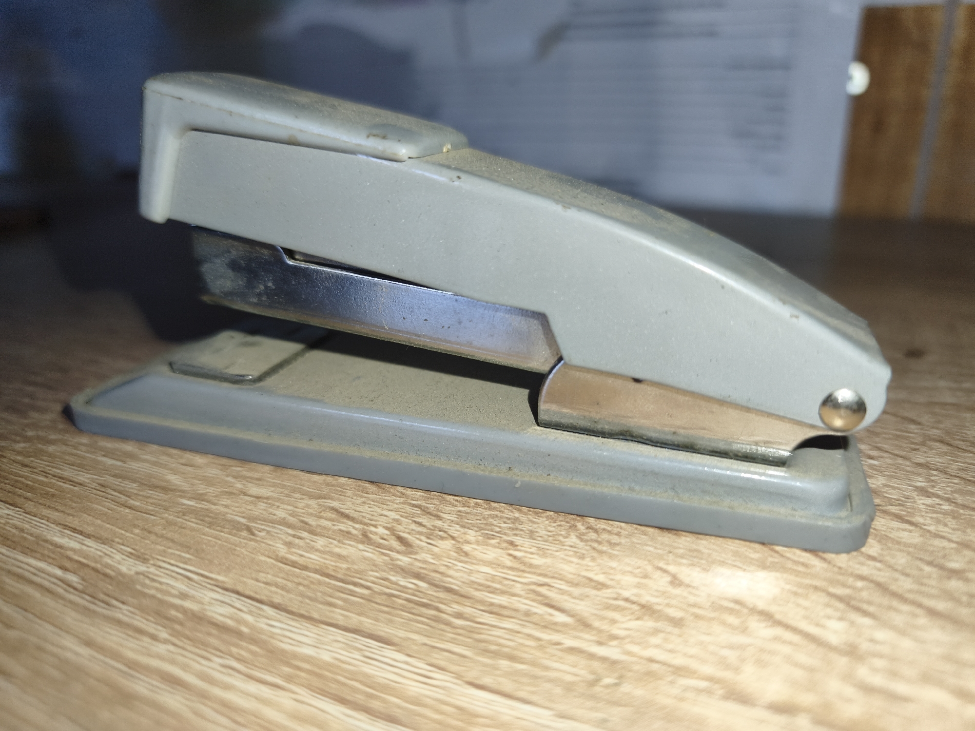The image shows a gray and silver stapler on a wooden surface. The stapler is dusty and appears to be old. The background is out of focus, but it looks like a desk with some papers on it. The stapler is positioned at an angle, with its front facing towards the right. The wooden surface has a light brown, wood grain pattern. There are no people or animals visible in the image. The lighting suggests an indoor setting, likely an office or home environment. The scene is static and shows a common office supply. The location in Гаранскі сельскі Савет, Belarus is not directly depicted or inferable from the visual elements present in the image.