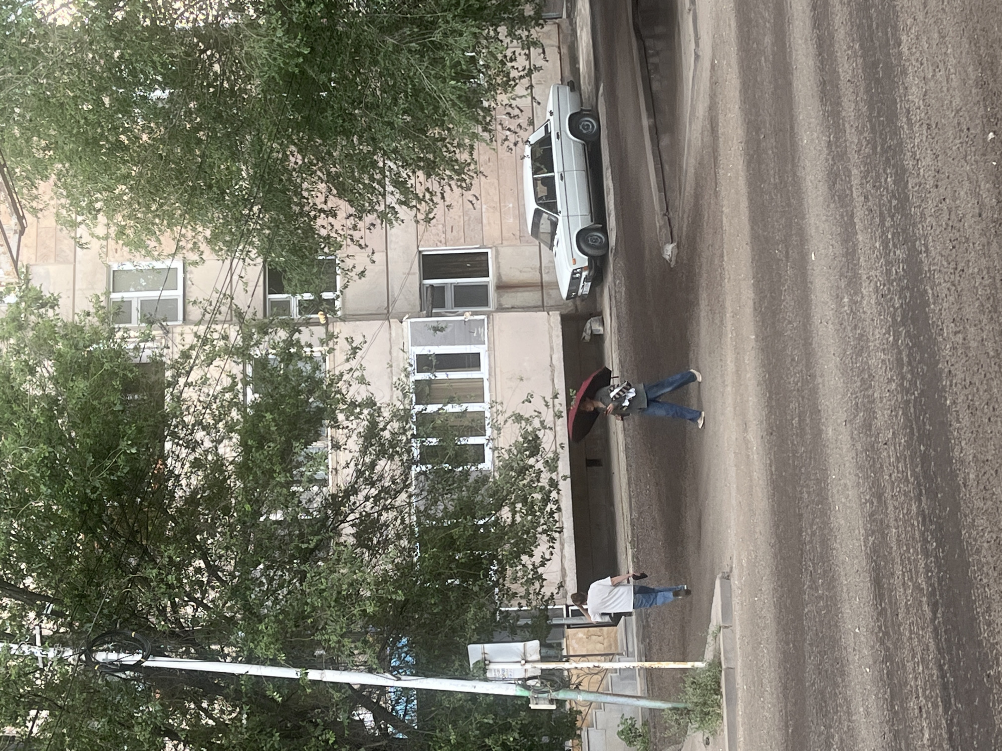 An outdoor, eye-level, medium shot captures a street scene in front of an apartment building. The building has a light-colored facade with multiple windows and balconies. In the foreground, a person walks away from the camera, holding what appears to be shoes in their hand. Further down the street, another person is walking while holding a red umbrella overhead. A white sedan is parked on the side of the road. Trees with green foliage flank the scene, obscuring parts of the building and sky. The street surface is dirt or gravel, with some areas appearing wet. A utility pole with wires is visible on the left side of the image. The lighting suggests it is daytime, possibly overcast.