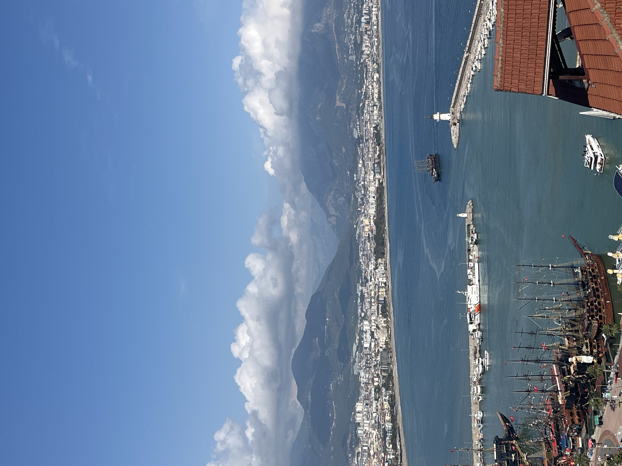 The image presents a partial view of Alanya, Turkey, likely taken from an elevated position. The scene is dominated by the blue of the Mediterranean Sea, which separates a coastline of densely packed buildings from mountainous terrain partially obscured by white clouds. The shoreline features a coastal city, with a visible line of buildings following the coast.
In the foreground, there is a harbor containing multiple docked vessels, including what appear to be decorative or tourist pirate ships with prominent masts and rigging. A couple of boats and a white lighthouse are seen near the pier. The corner of a red-tiled roof is visible in the upper right corner.
The sky to the left of the mountains is a clear blue, contrasting with the overcast and hazy conditions around the mountains.