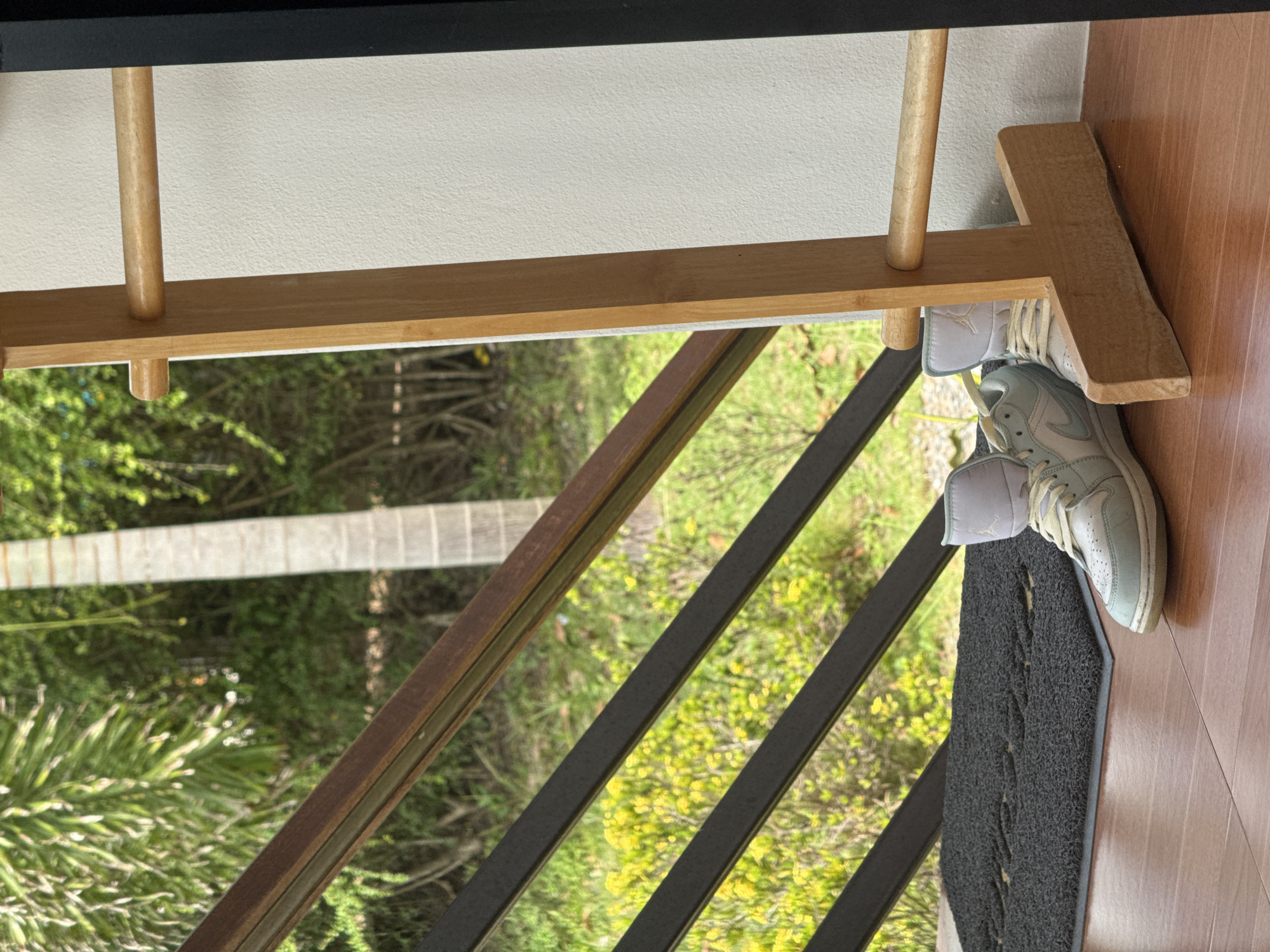 A pair of light blue and white high-top sneakers rests on a wooden floor in front of a doormat. The sneakers are positioned next to a light-colored wall and a wooden rack. In the background, there is a railing with dark metal bars and a brown-toned wooden top. Beyond the railing, lush green foliage and a palm tree trunk are visible, suggesting an outdoor or semi-outdoor setting, potentially in a tropical location like Ko Samui District, Thailand. The lighting appears natural, indicating it is daytime.