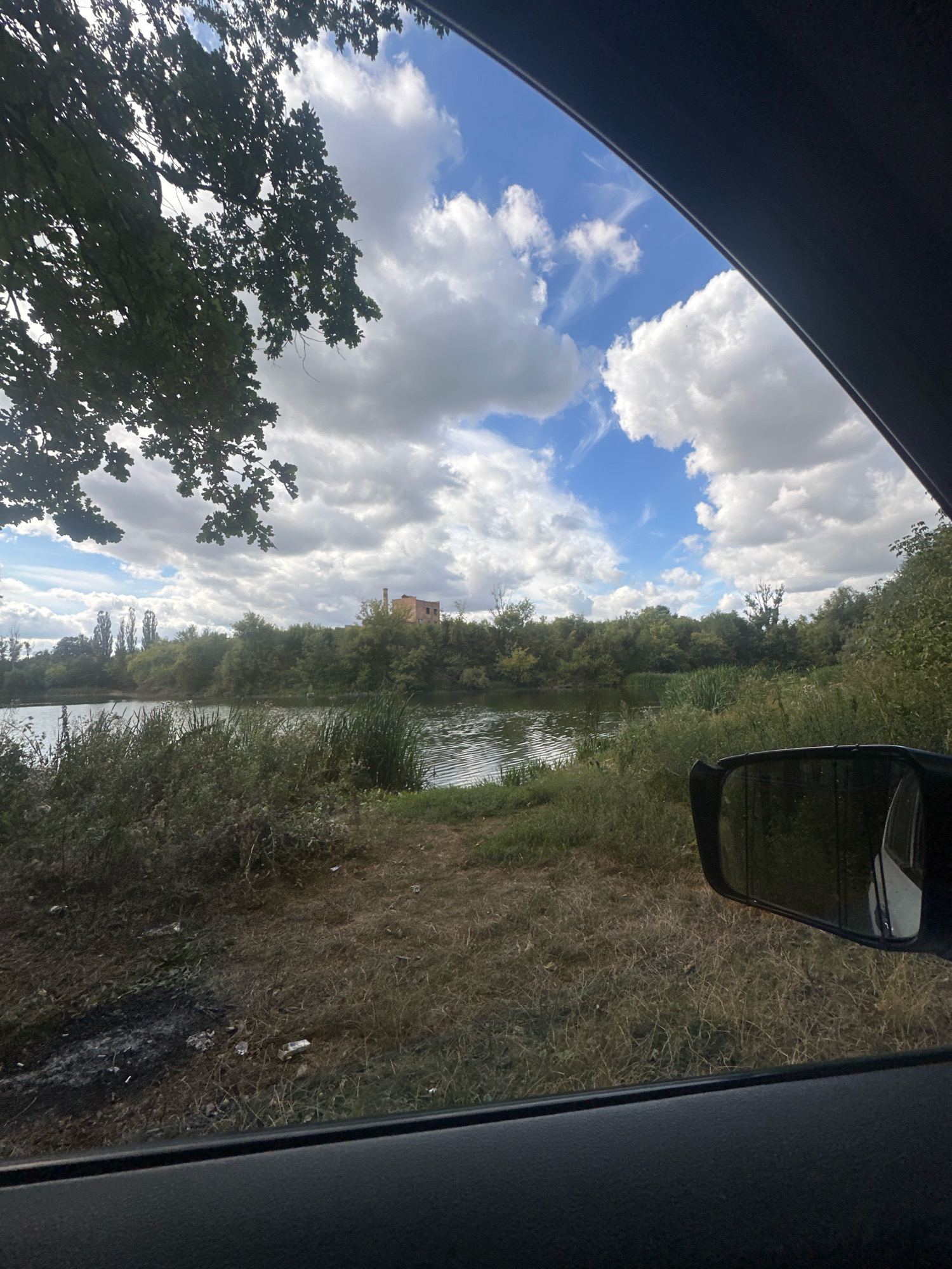 The image shows a view from inside a vehicle, looking out towards a body of water. A large tree with green leaves overhangs the left side of the frame. The sky is blue with white, puffy clouds and some gray clouds. Across the water, there is a dense line of green trees and vegetation. In the distance, on top of a hill, is a small, reddish-brown building. A side-view mirror of the vehicle is visible in the lower right corner of the image. The foreground consists of dry, brown grass and some scattered debris. The scene appears to be a natural area, possibly a park or a rural landscape, with the building suggesting some human presence in the vicinity. The location is identified as Makiv, Ukraine.