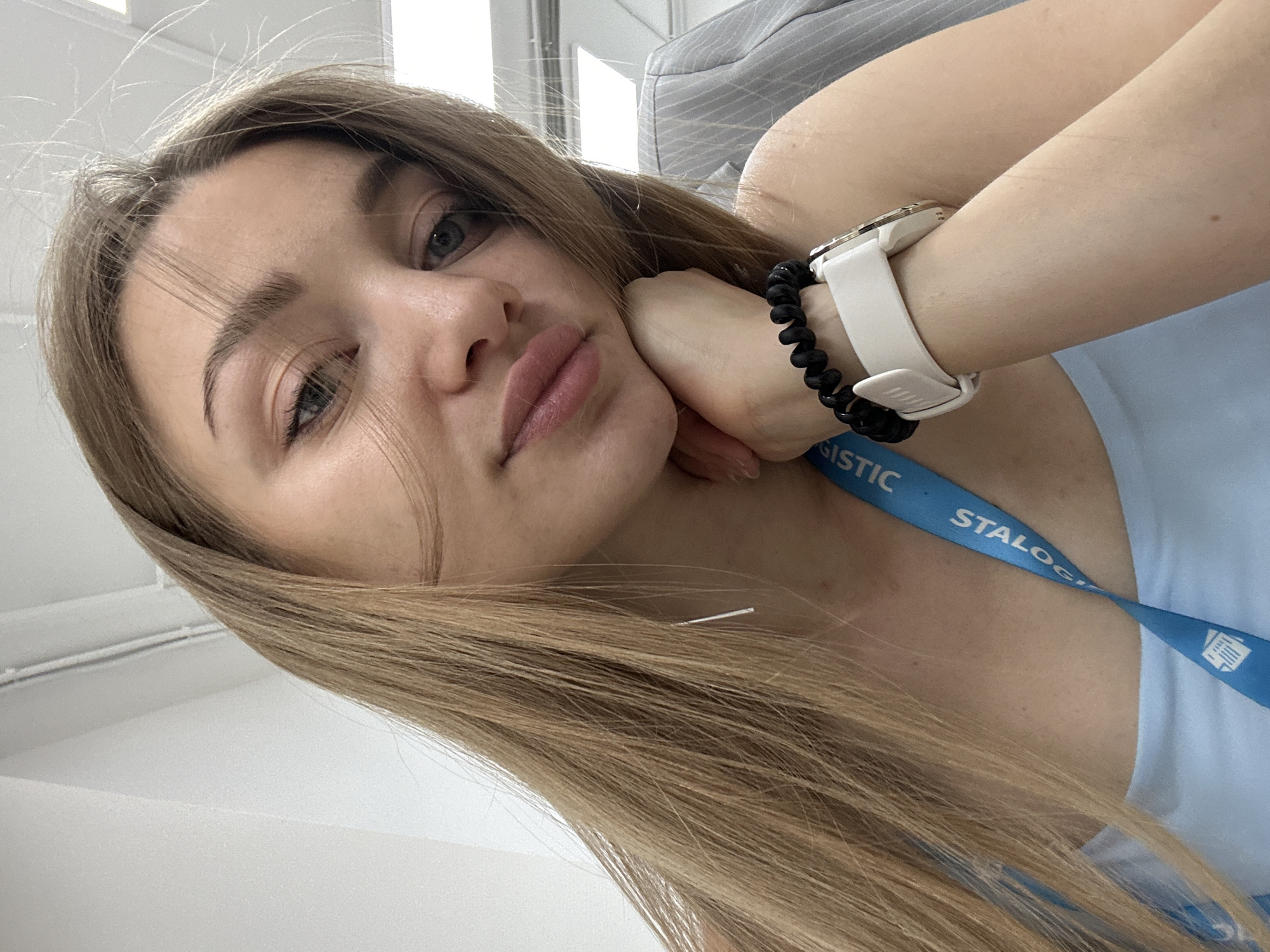 A woman with long, straight, light brown hair is posing for a selfie. She is leaning her head on her left hand, which is adorned with a white smartwatch and a black coiled bracelet. She has fair skin, light eyebrows, and light-colored eyes. She is wearing a light blue top and a blue lanyard with white lettering partially visible, reading "STALOGISTIC". The background includes a light-colored wall, a striped object that appears to be a part of a chair or clothing, and a light fixture in the upper part of the frame. The location is undetermined, however based on text, it is likely within Kazan, Russia.