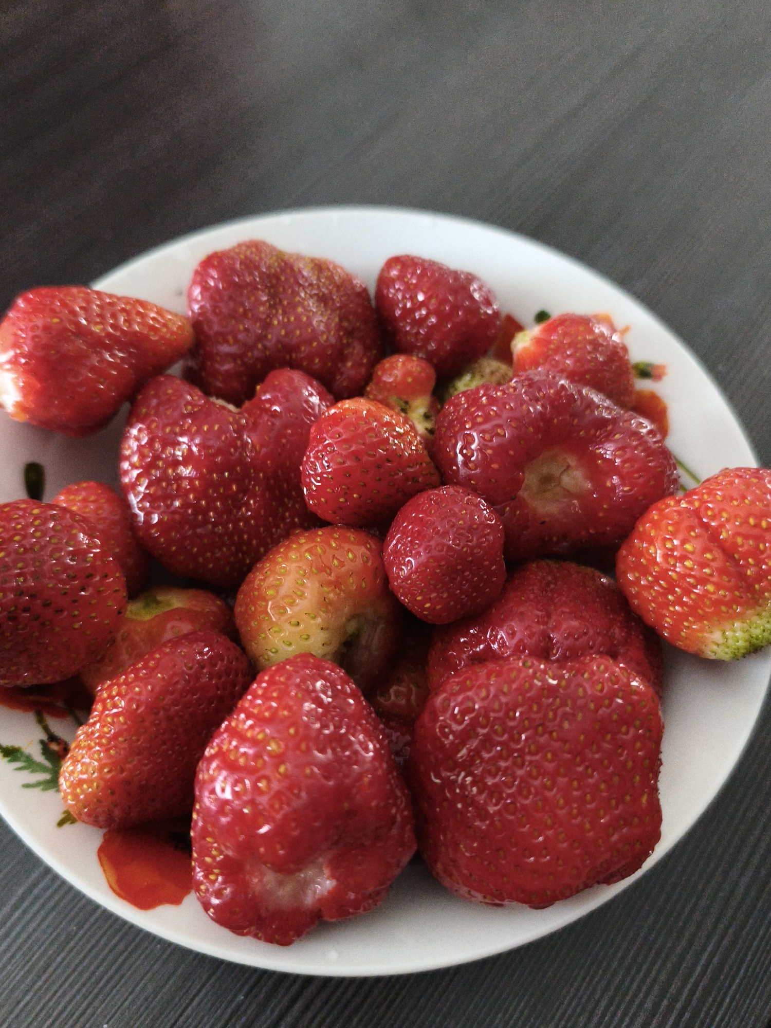 A close-up, overhead shot features a white, circular plate filled with a variety of fresh strawberries. The strawberries are predominantly bright red, with some exhibiting lighter red and yellow hues. A few strawberries show visible green tips, indicating they are nearing ripeness or are just past their peak. The plate has a subtle floral pattern along its rim. The strawberries are arranged densely, with some overlapping. The background consists of a dark, textured surface, possibly a wooden table, with a subtle sheen. The lighting is even, highlighting the glossy texture of the strawberries. This image was taken indoors.