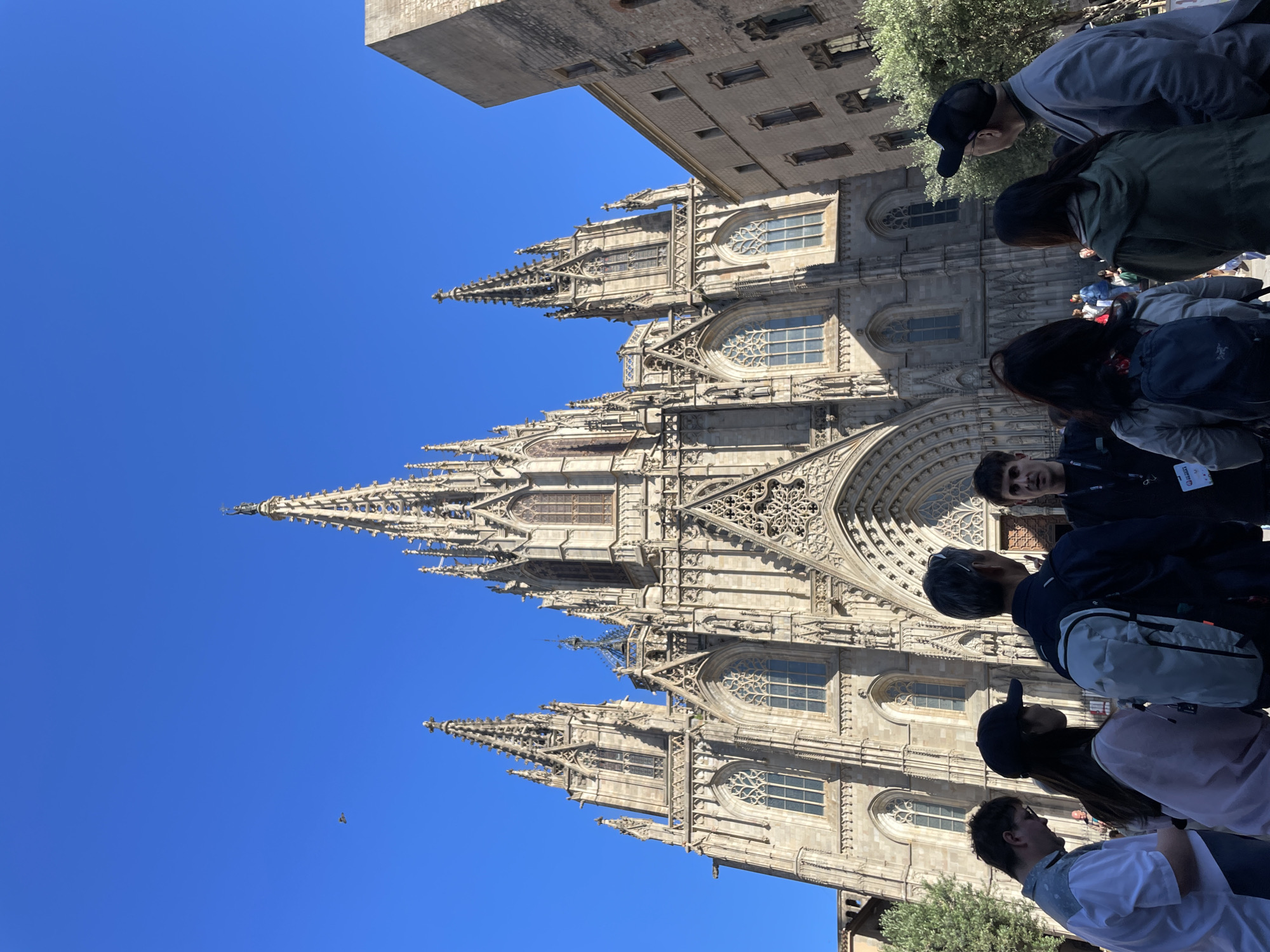 The image shows the Barcelona Cathedral (Cathedral of the Holy Cross and Saint Eulalia), a Gothic cathedral in Barcelona, Spain. The facade of the cathedral is intricately detailed, with spires and ornate windows. A group of people, possibly tourists, are standing in front of the cathedral. The sky is clear and blue. The cathedral and building behind it are constructed of grey stone. The light suggests it is during the daytime.