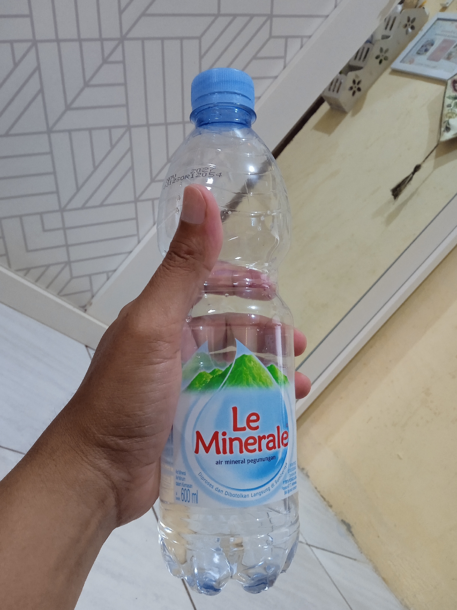 A person's hand holds a clear plastic bottle of "Le Minerale" mineral water. The bottle has a blue cap and the label features green mountains and the product name in red. The text on the label includes "air mineral pegunungan" and "600 ml". The bottle's expiration date on top is NOV 2027. The background includes a geometric patterned wall and a beige surface with decorative elements. The location is Cangkring, Indonesia.
