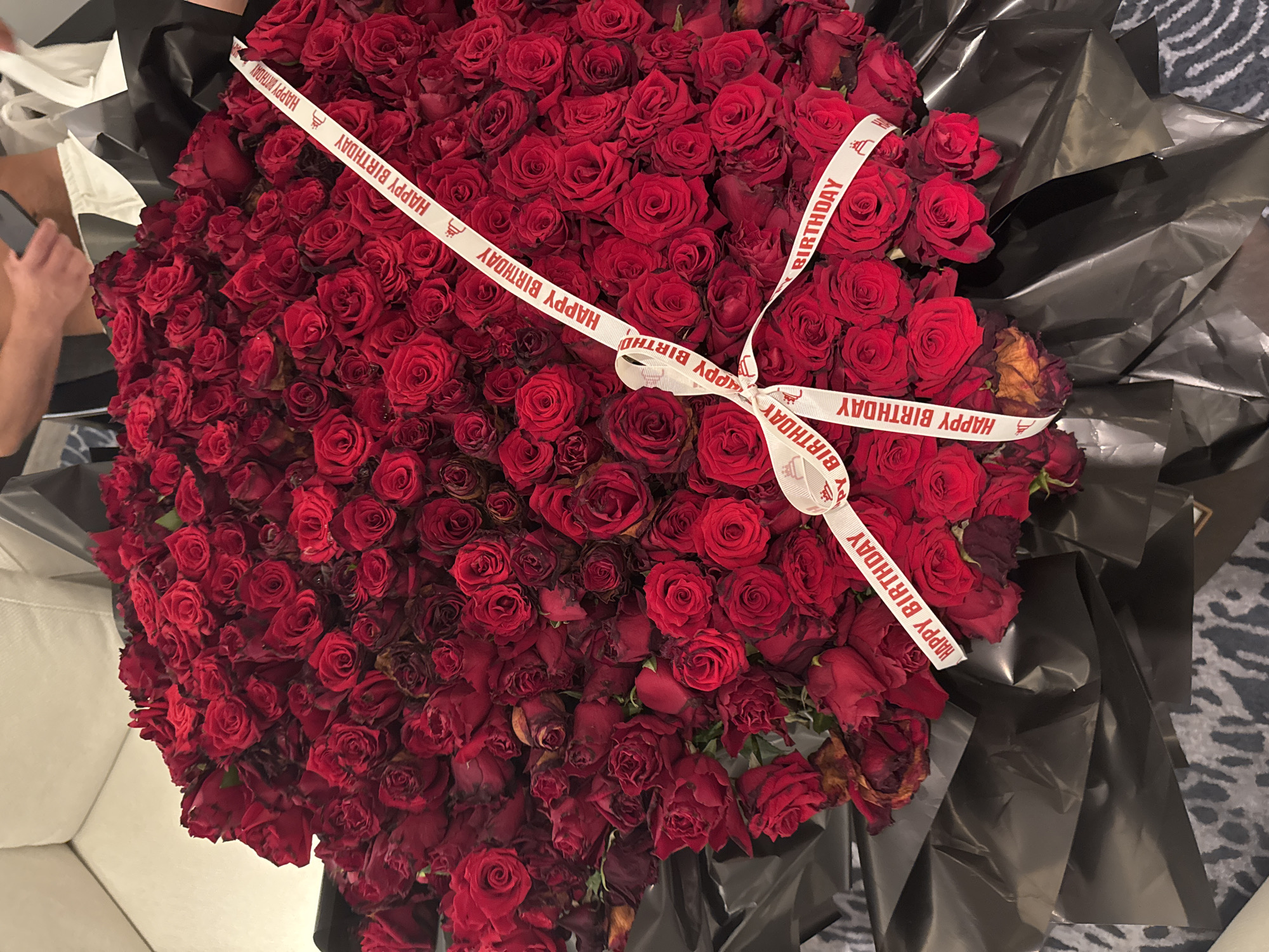 A top-down shot reveals a large bouquet of red roses wrapped in black plastic sheeting. A white ribbon with the words "HAPPY BIRTHDAY" printed in red runs diagonally across the bouquet and is tied in a bow in the center. A person is visible in the upper left corner holding a phone. The bouquet is sitting on either a sofa or a floor. The location is not stated.