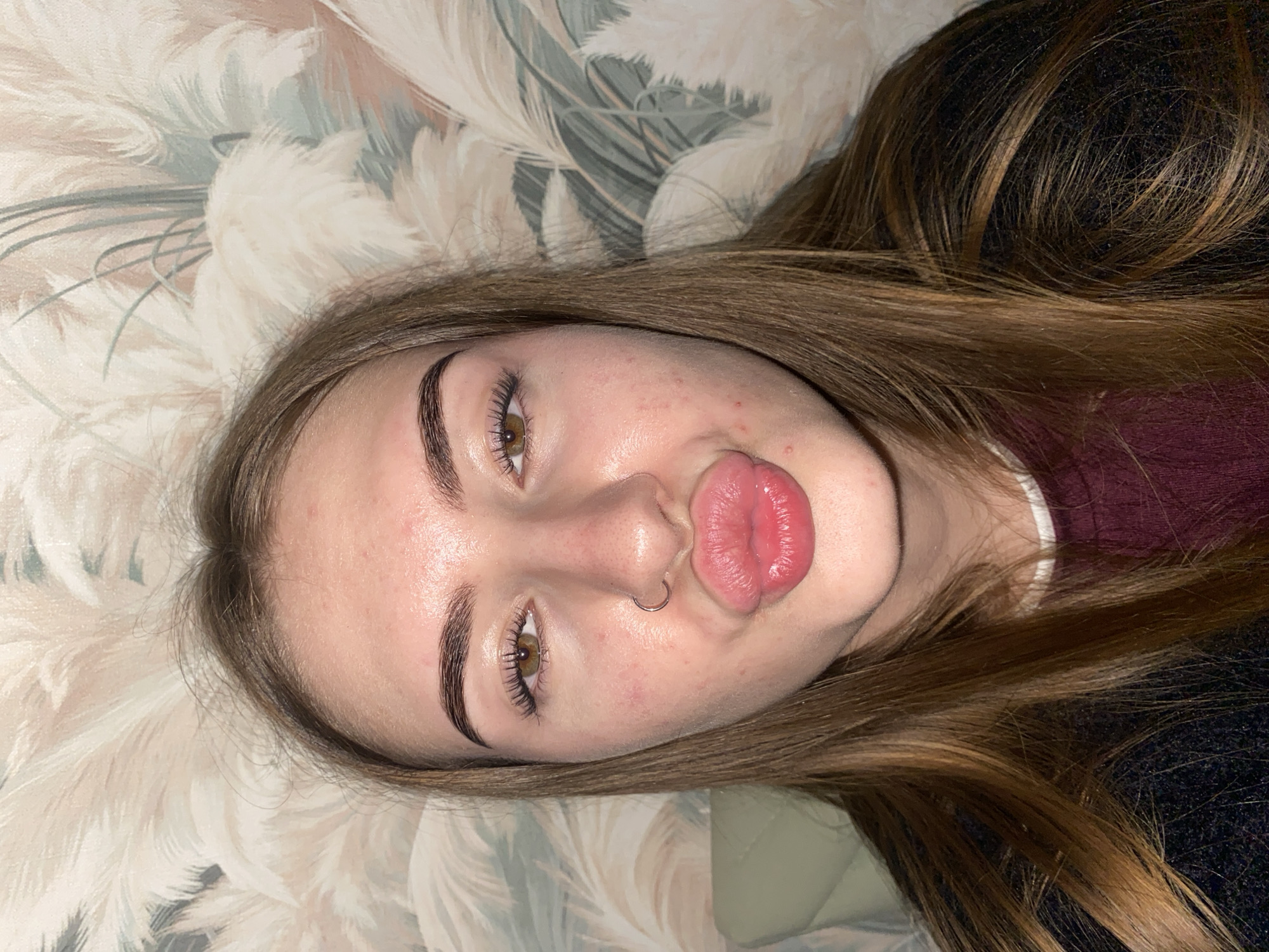 A close-up, top-down shot captures a young individual lying down, with their head tilted to the right. The subject has medium-long, light brown hair with subtle highlights, which is spread out around their head. They have defined, dark eyebrows and long, dark eyelashes framing light brown or hazel eyes. A small silver hoop piercing is visible on their left nostril. Their lips are pursed into a "kissing" or "duck face" expression, appearing full and a glossy pink shade. Their skin is fair with a few minor blemishes, particularly on their forehead and cheek.
A maroon top with a visible white collar underneath is partially visible on the right side of the frame, beneath their hair. Another darker fabric, possibly black or dark blue and textured, is seen further down. The background consists of a light-colored pattern featuring flowing, feathery or leafy shapes in shades of beige, light brown, and pale green/grey, suggesting wallpaper or bedding. The lighting appears to be indoor, possibly artificial, illuminating the subject's face evenly. The overall mood conveyed by the expression is playful and casual. There is no visible text in the image.