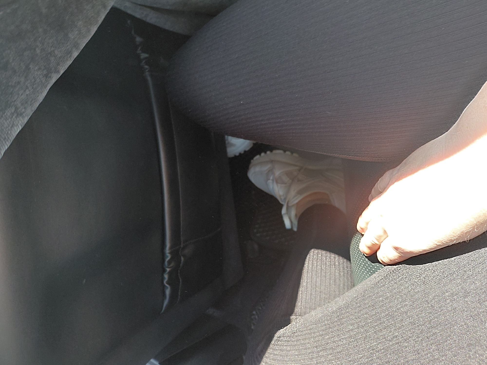 The image captures a close-up view of a person's legs and hand inside a vehicle. The perspective is from the back seat, looking towards the front. In the foreground, on the left, is a textured dark gray fabric, likely part of the car's interior or the person's clothing. To the right, a person's hand with visible fingers is resting on this fabric. Further into the car, a white sneaker is visible, positioned near the pedals on the floor. The seat in front is a dark, possibly black, material, with a pocket on its back. The overall lighting suggests it is daytime, with sunlight casting shadows. The scene is devoid of any discernible urban context or specific landmarks of Chulym, Russia, making its location within the city impossible to determine from the image alone.