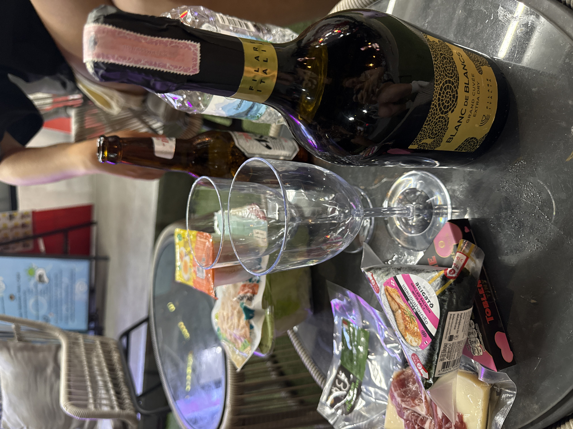 This image captures a close-up, top-down view of a table laid out with food and drinks, likely for a casual gathering. The scene appears to be indoors, possibly a dining area or a patio, with a hint of natural light suggesting daytime.
The primary subjects are a bottle of sparkling wine, identified by the label "BLANC DE BLANCS GRAND CUVÉE EXTRA DRY FILARE ITALIA," and two empty wine glasses. Beside the wine bottle, there's a brown glass bottle, partially obscured, possibly a beer.
Arranged on the table are various food items, some still in their packaging. Notable among these are individually wrapped rice balls with seaweed, one clearly labeled "ARIGATÓ GRILLED SALMON WITH SALT" in both English and Thai script. There's also a package of thinly sliced cured meat, likely ham or prosciutto, and a wedge of cheese. Smaller packets of what appear to be snacks or condiments are also visible. A red and white package labeled "TOBLER" suggests chocolate is also part of the spread.
The overall impression is one of an informal get-together or picnic, with a selection of wine, drinks, and convenient, ready-to-eat food items. The lighting is somewhat dim with reflections on the table surface, creating a slightly glossy effect. The orientation of the image is also unconventional, appearing rotated. The specific text on the bottles and food packaging provides detailed information about the products. The setting appears to be in Baan Bo Phut, Thailand, as indicated by the Thai script on the food packaging.
