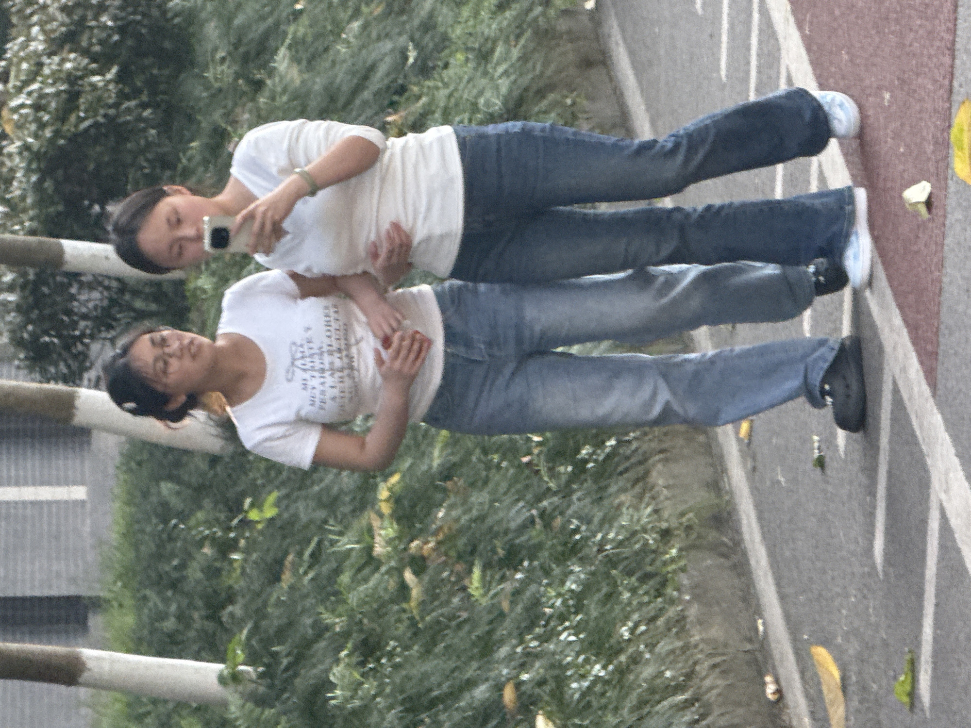 Two women stand on a pedestrian walkway alongside greenery and bare-trunked trees. The women are dressed in white t-shirts and jeans; one woman holds a cell phone up as if taking a photo. The walkway is painted with white lines and sits next to a red running track. Fallen leaves are scattered across the pathway. The location is suggestive of Jiangjin, China, given the surrounding foliage and pavement style.
