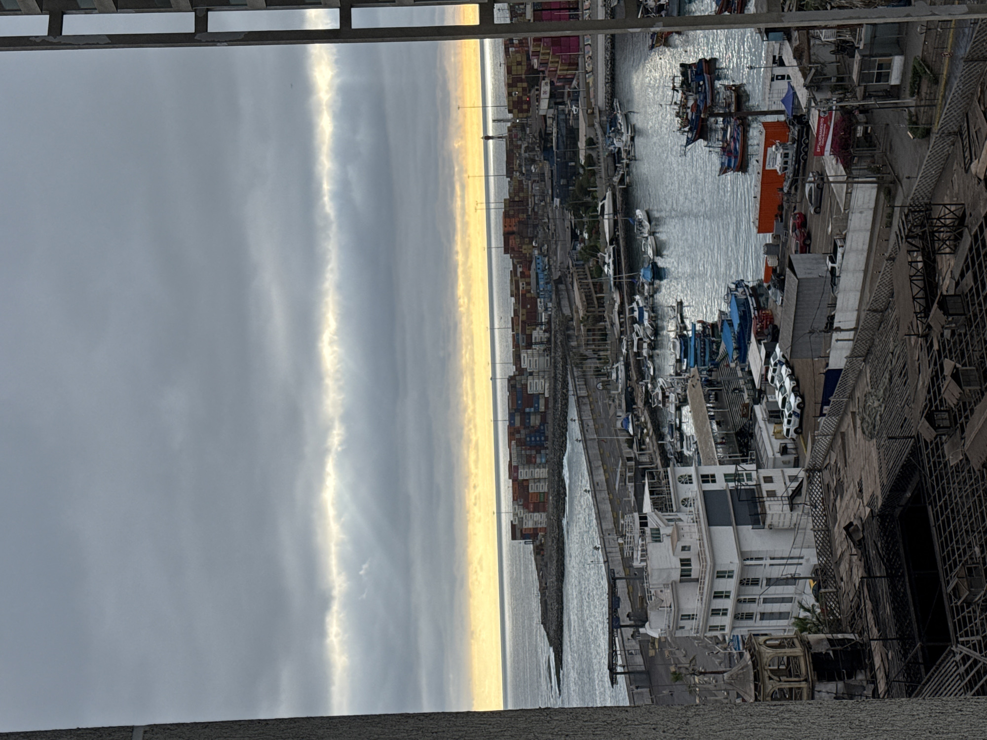 The image depicts a wide-angle view of a port in Iquique, Chile, under a cloudy sky with rays of sunlight breaking through. In the foreground, the roof of a partially destroyed building is visible, with exposed wooden beams and debris. To the left, a weathered, ornate structure with a domed roof stands. Adjacent to this is a tall, white modern building with multiple floors and windows. Beyond these structures, a marina is filled with numerous boats of various sizes, many of them small fishing vessels. A paved area with parked cars, including several white police vehicles, runs alongside the marina. Further into the scene, a long pier extends into the water, lined with shipping containers of different colors. The ocean stretches out to the horizon, meeting the sky where the sun is setting or rising, casting a warm glow through the clouds. The overall impression is one of a working port, with elements of urban development and the aftermath of some structural damage.
