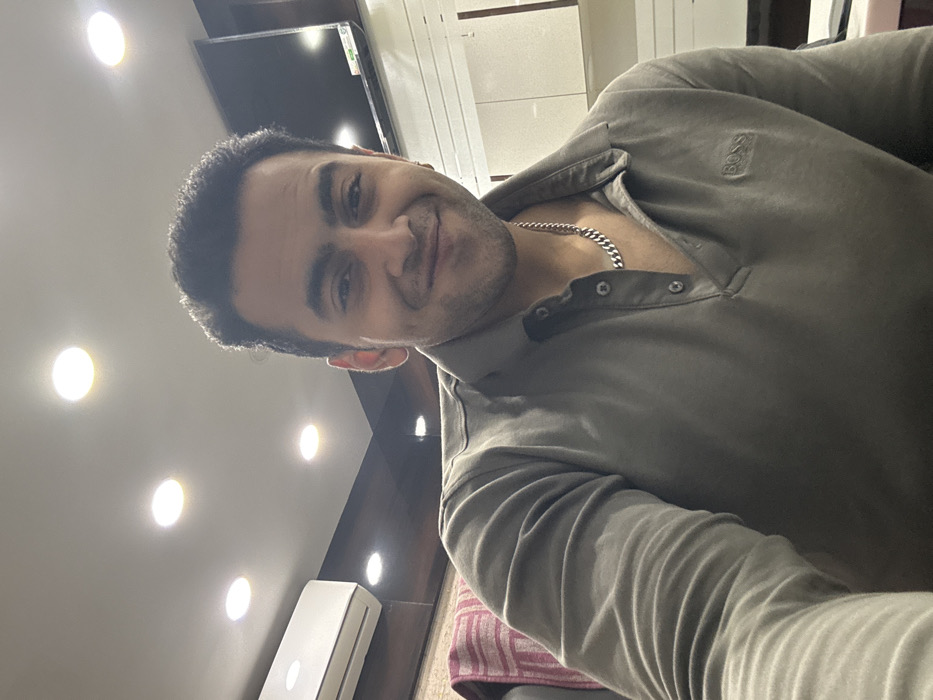 The image features a man taking a selfie indoors. He has short dark hair, dark eyes, and a subtle smile on his face, looking directly at the camera. He is wearing a long-sleeved polo shirt in a grey or olive green color. A silver-colored chain is visible around his neck, and a small "BOSS" logo is embroidered on the left chest of his shirt.
The background indicates an indoor setting, possibly a modern living room or office. The ceiling is equipped with several bright, circular recessed lights. A flat-screen television is mounted on a wall in the upper left background, below which a white split air conditioning unit is installed. Parts of a dark accent wall, possibly made of wood or dark paneling, are visible, contrasting with lighter-colored walls. In the lower right corner, a striped fabric with pink/red and white patterns, likely a cushion or blanket, can be seen. The scene is brightly lit by artificial overhead lighting. The image was taken in Kolkata, India.